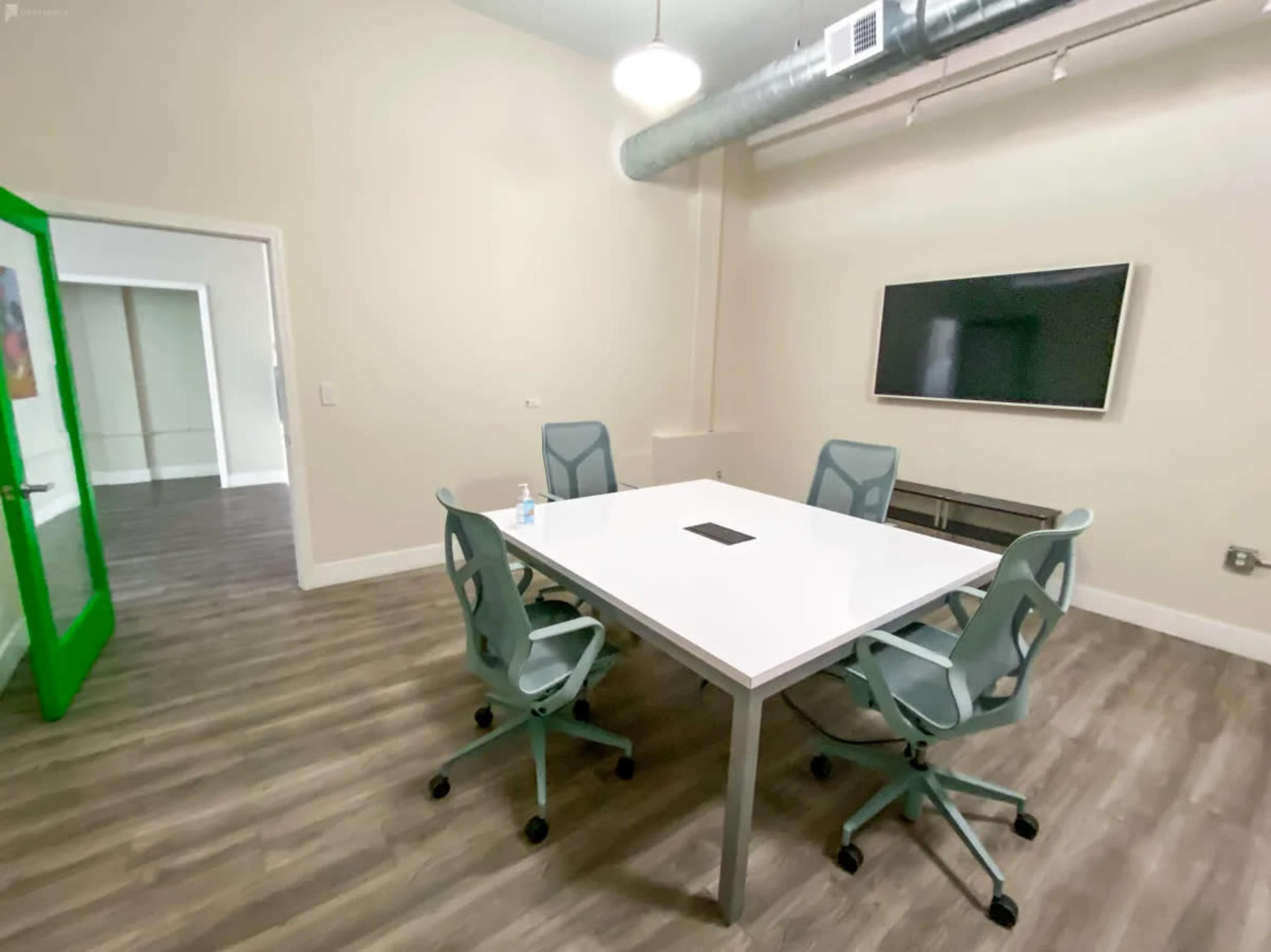 A conference room features a large white table surrounded by four light gray chairs, with a television mounted on the wall and a green door leading to another room.