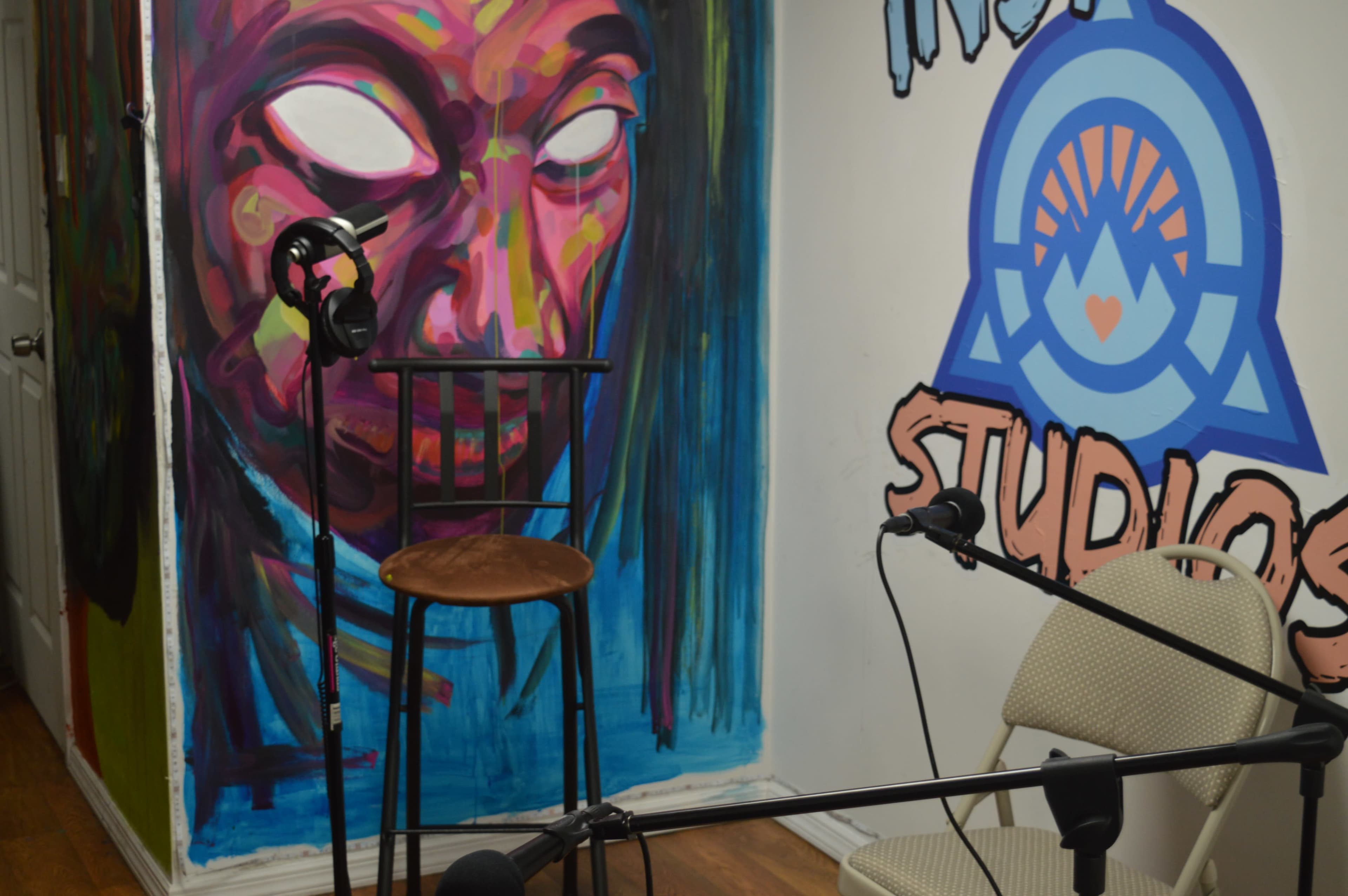 A recording studio corner with a colorful mural, a tall barstool, and microphones positioned on the floor.