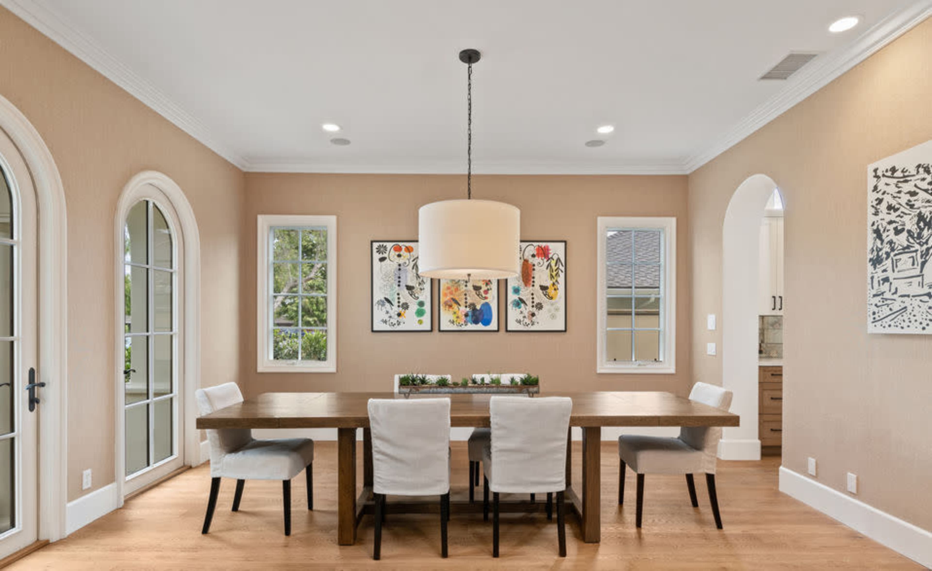 A spacious dining room features a wooden table surrounded by four chairs, with framed artwork on the walls and large windows allowing natural light.
