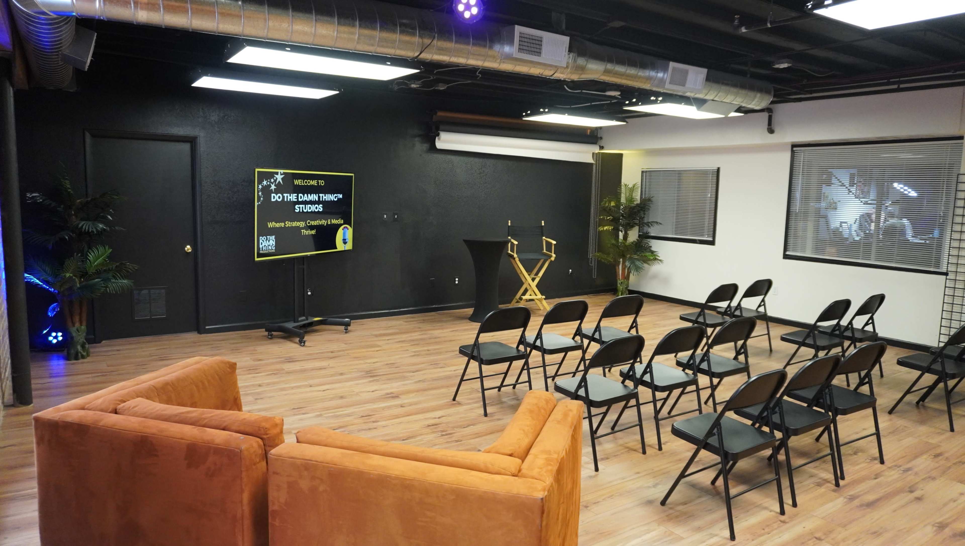 A minimalist studio space set up with rows of black chairs facing a monitor and two director's chairs beside a tall podium.