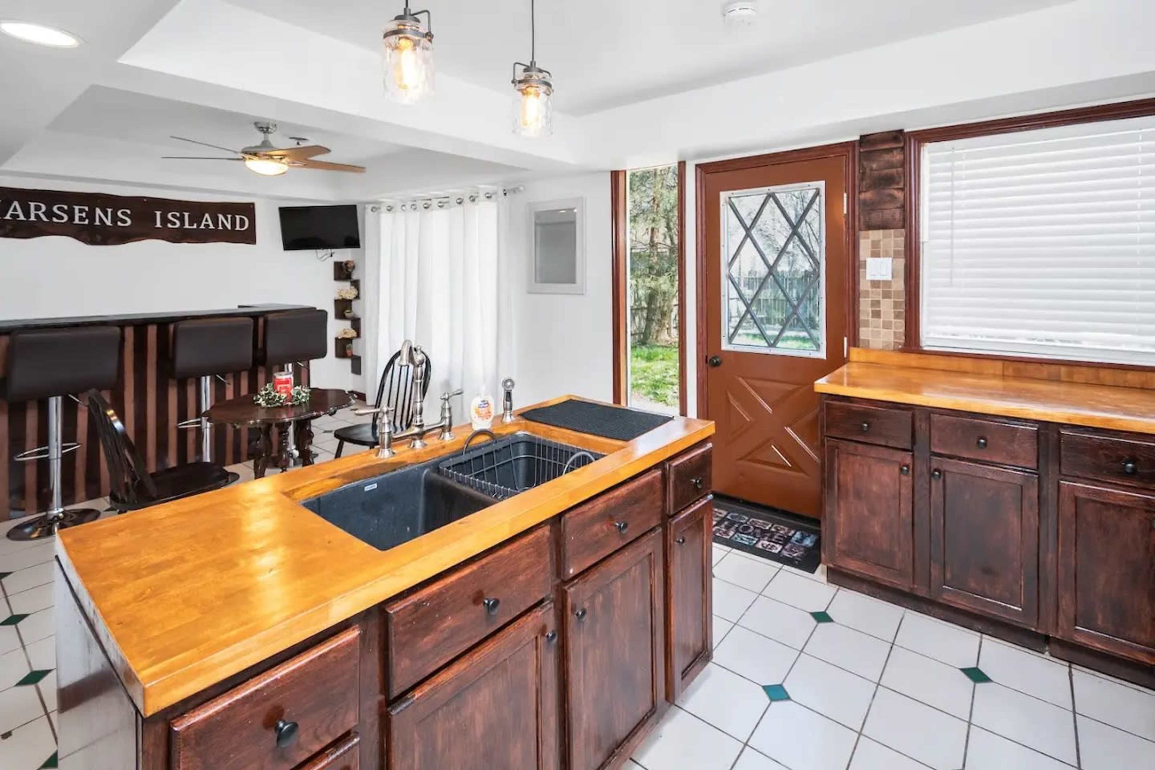 A kitchen with wooden cabinets, a double sink on a central island, and a door leading outside, framed by large windows.