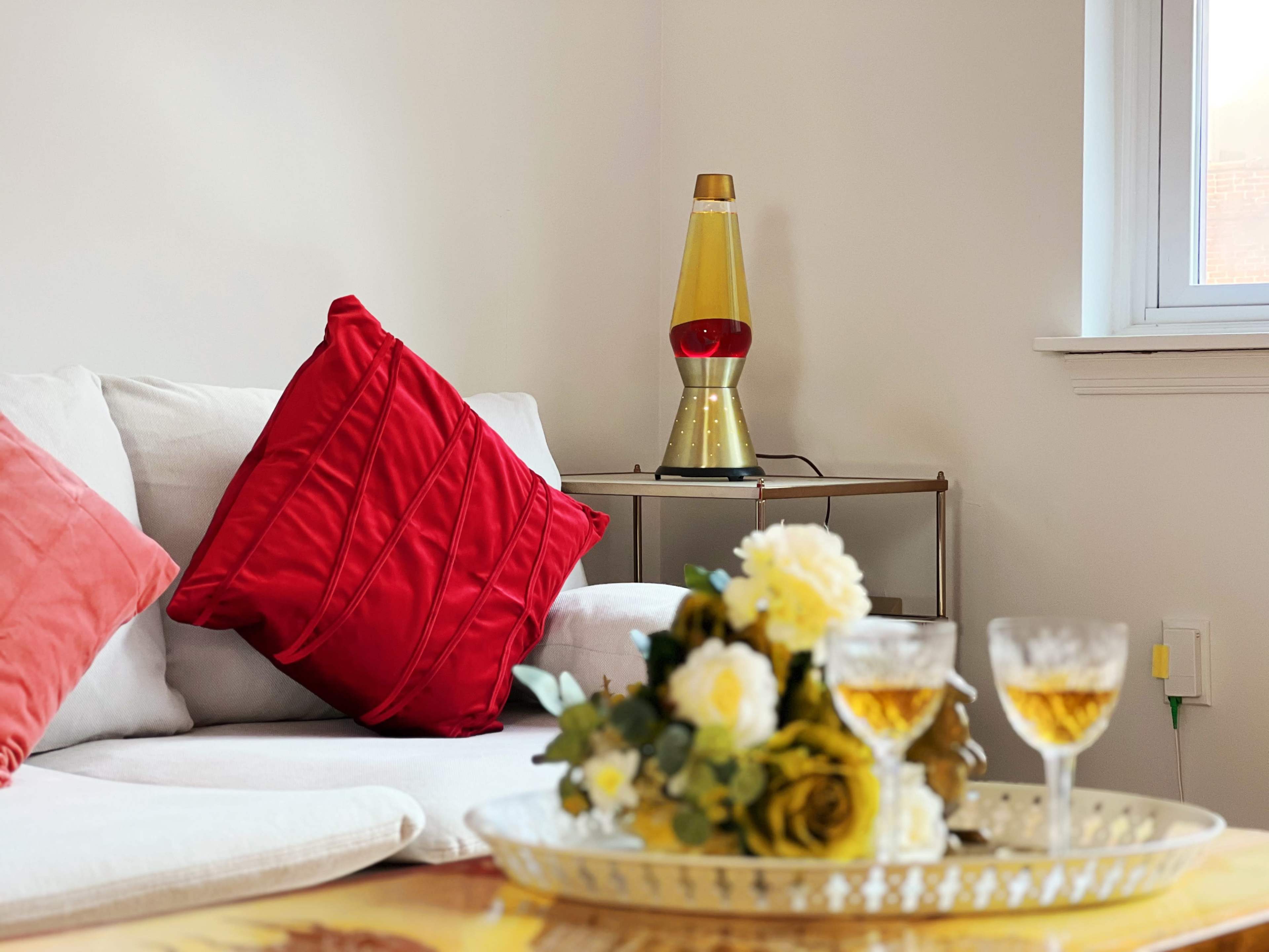 A cozy living room features a light-colored sofa with red pillows, a decorative tray with flowers and glasses, and a lava lamp on a side table.
