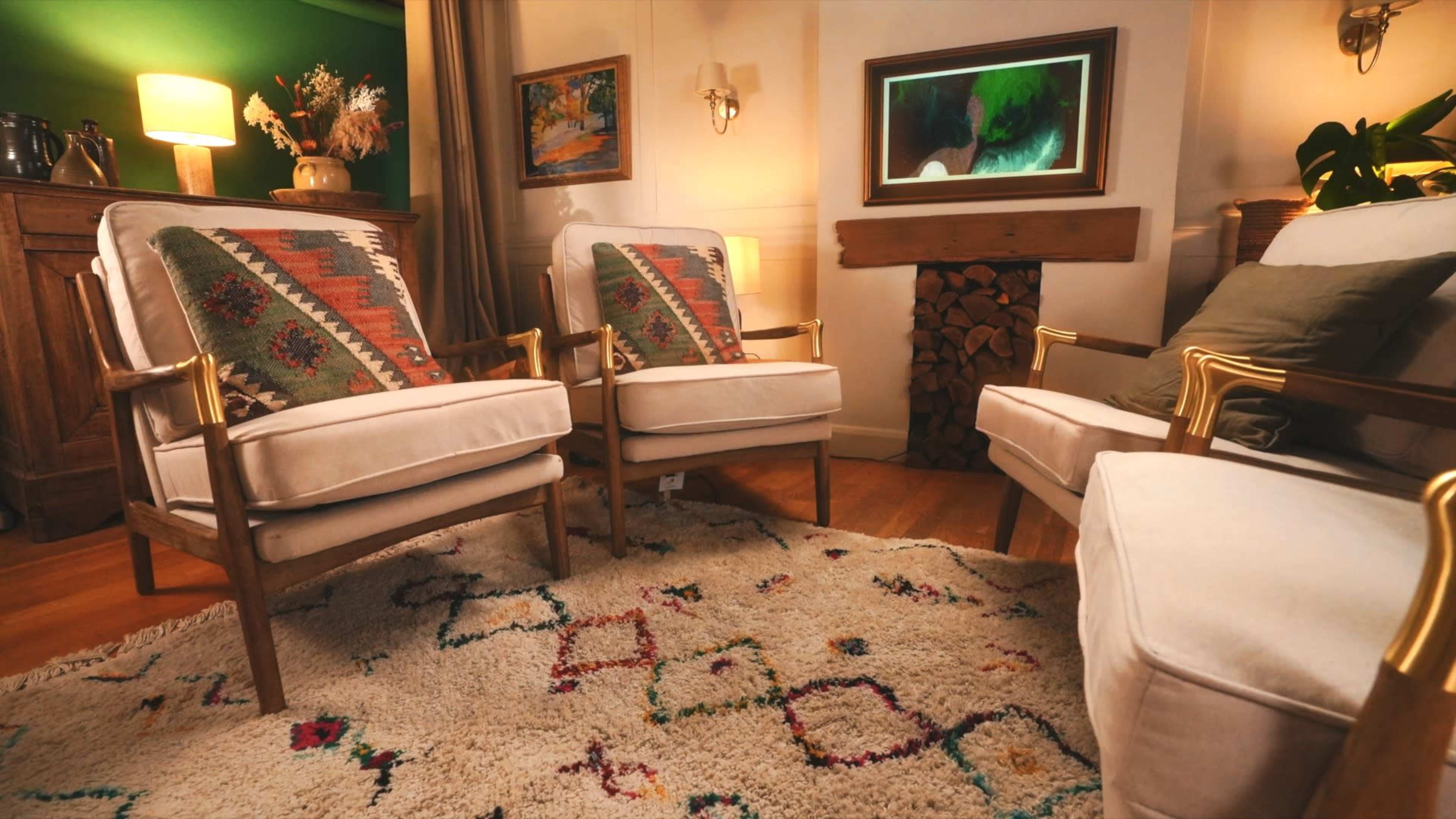 The image shows a cozy living room with two upholstered armchairs facing each other, a decorative rug on the floor, and a wooden accent wall with stacked wood.