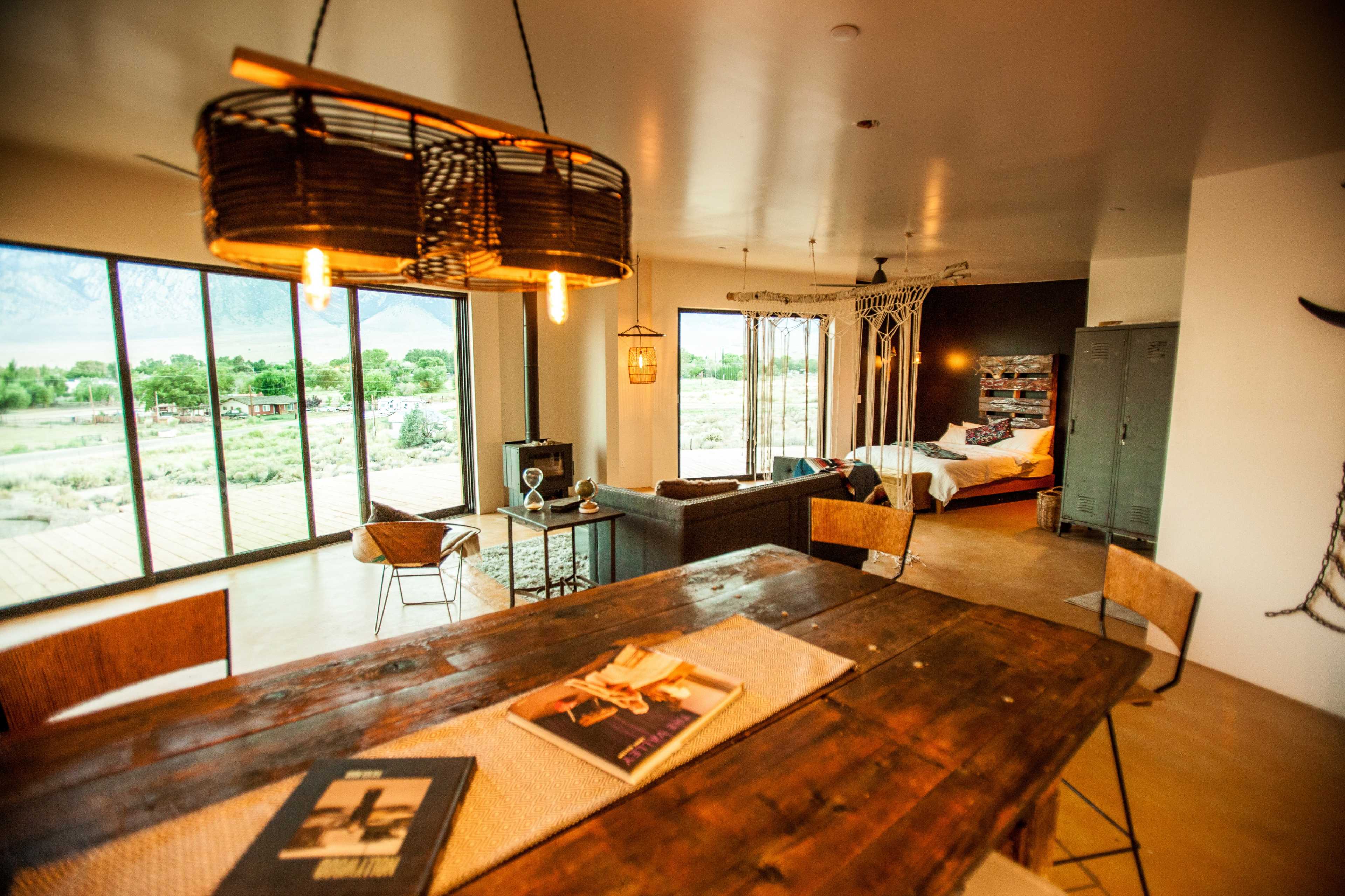 The image shows a modern interior space featuring a dining table, seating area, and a bed in the background with large windows offering a view of the outdoors.