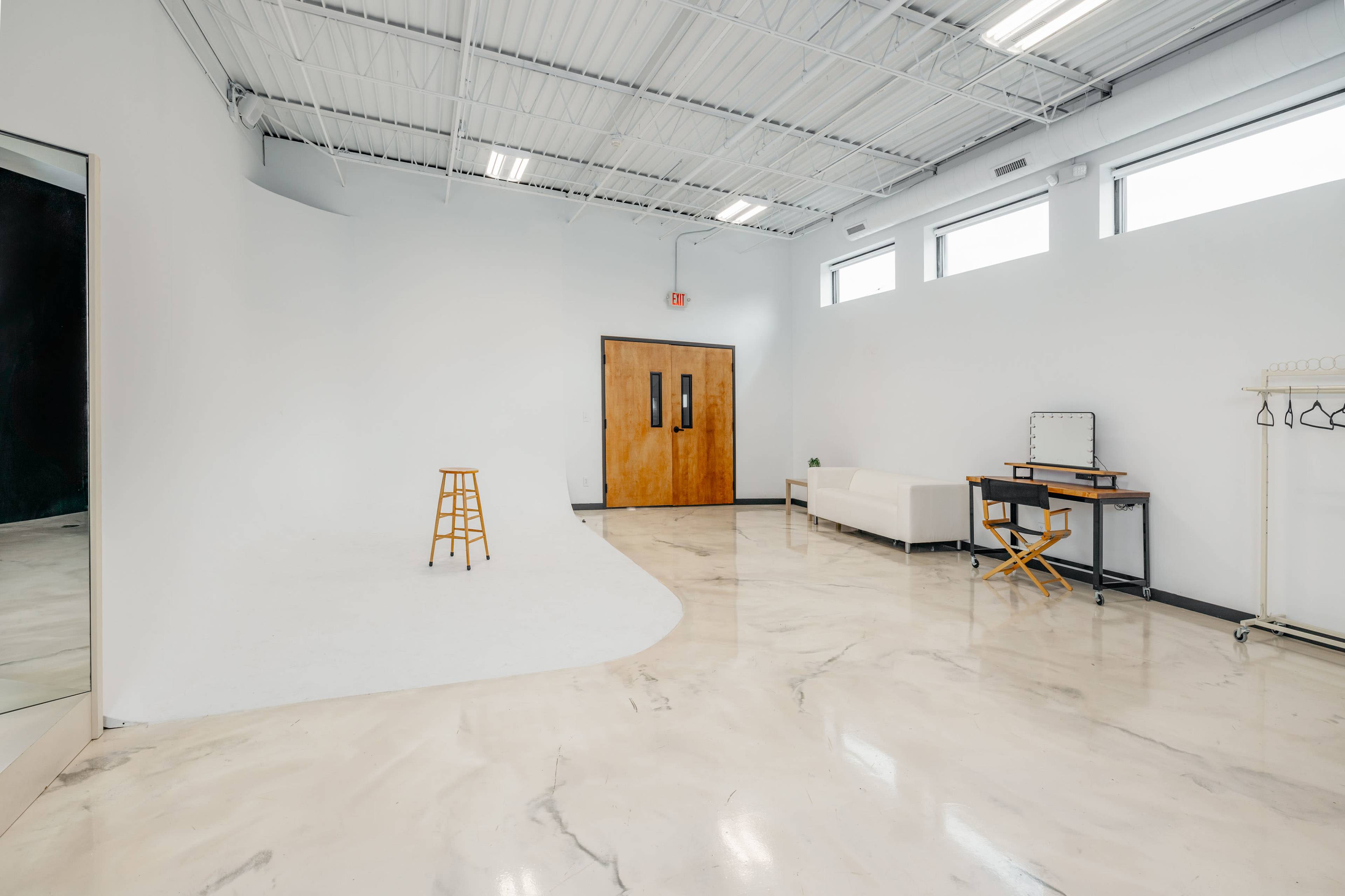 An empty studio space with a white curved backdrop, a wooden stool, a couch, a desk with a mirror, and windows letting in natural light.