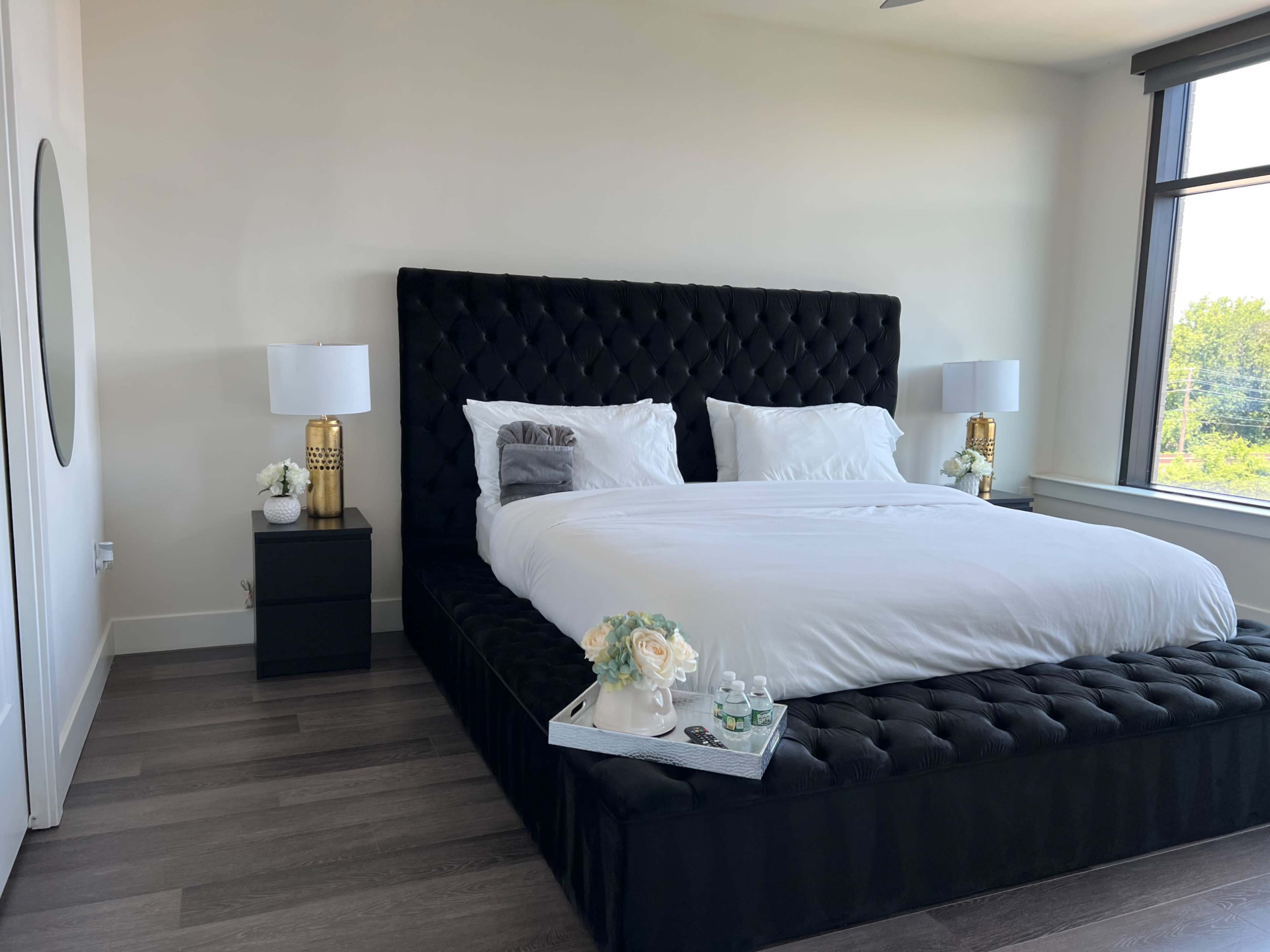 A modern bedroom with a large tufted black bed, two bedside lamps on either side, and a small decorative tray with flowers on the bed.