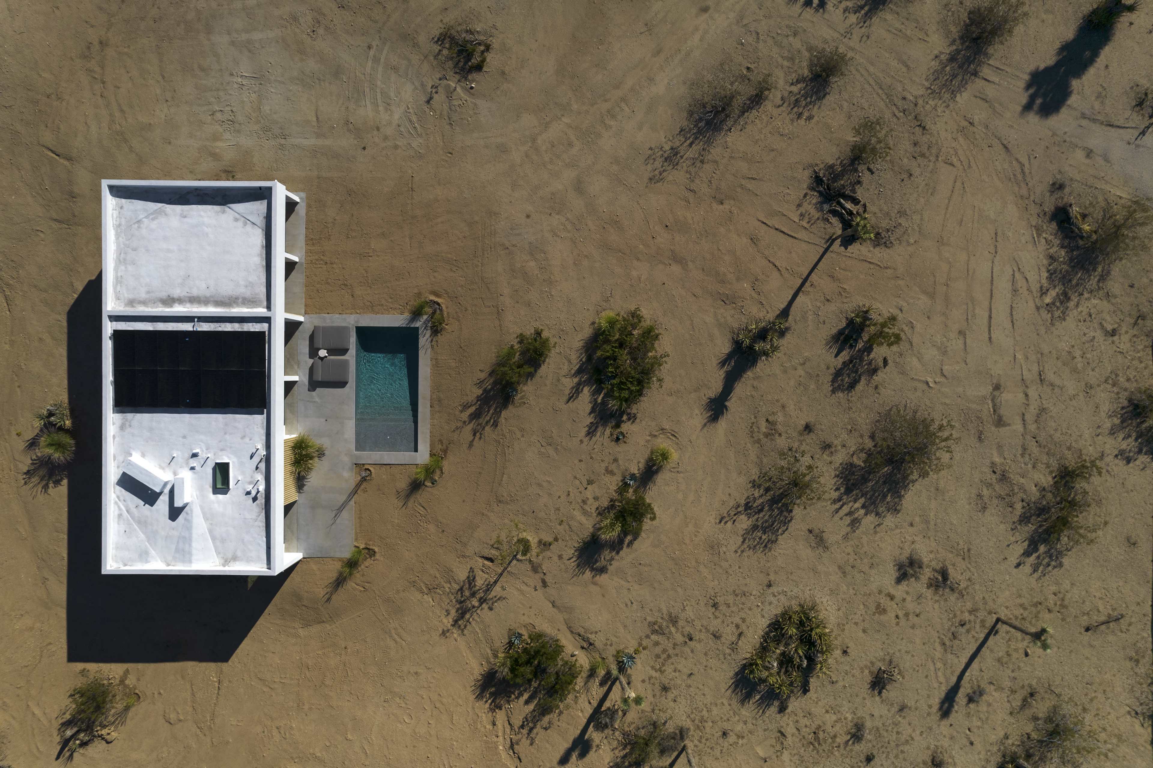 A modern, minimalist house with a pool is situated in a barren desert landscape surrounded by sparse vegetation.