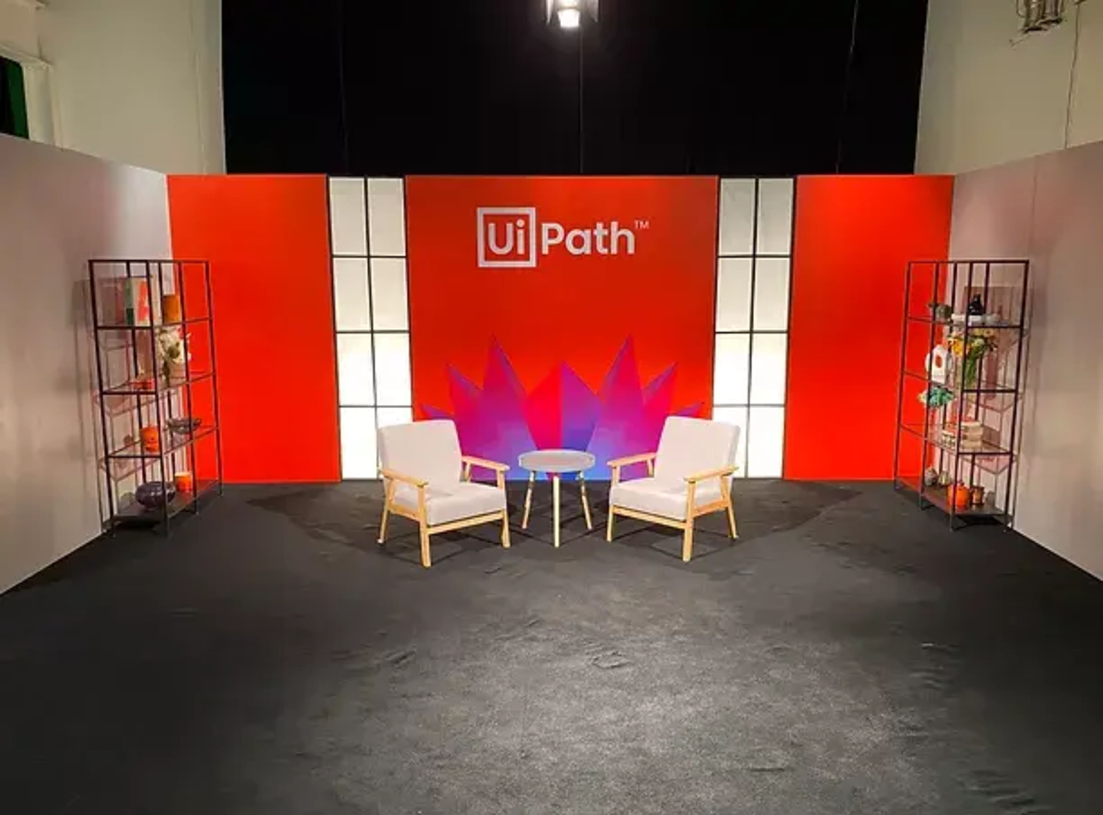 A stage setup with two white chairs and a round table in front of a vibrant backdrop featuring the UiPath logo and decorative elements.