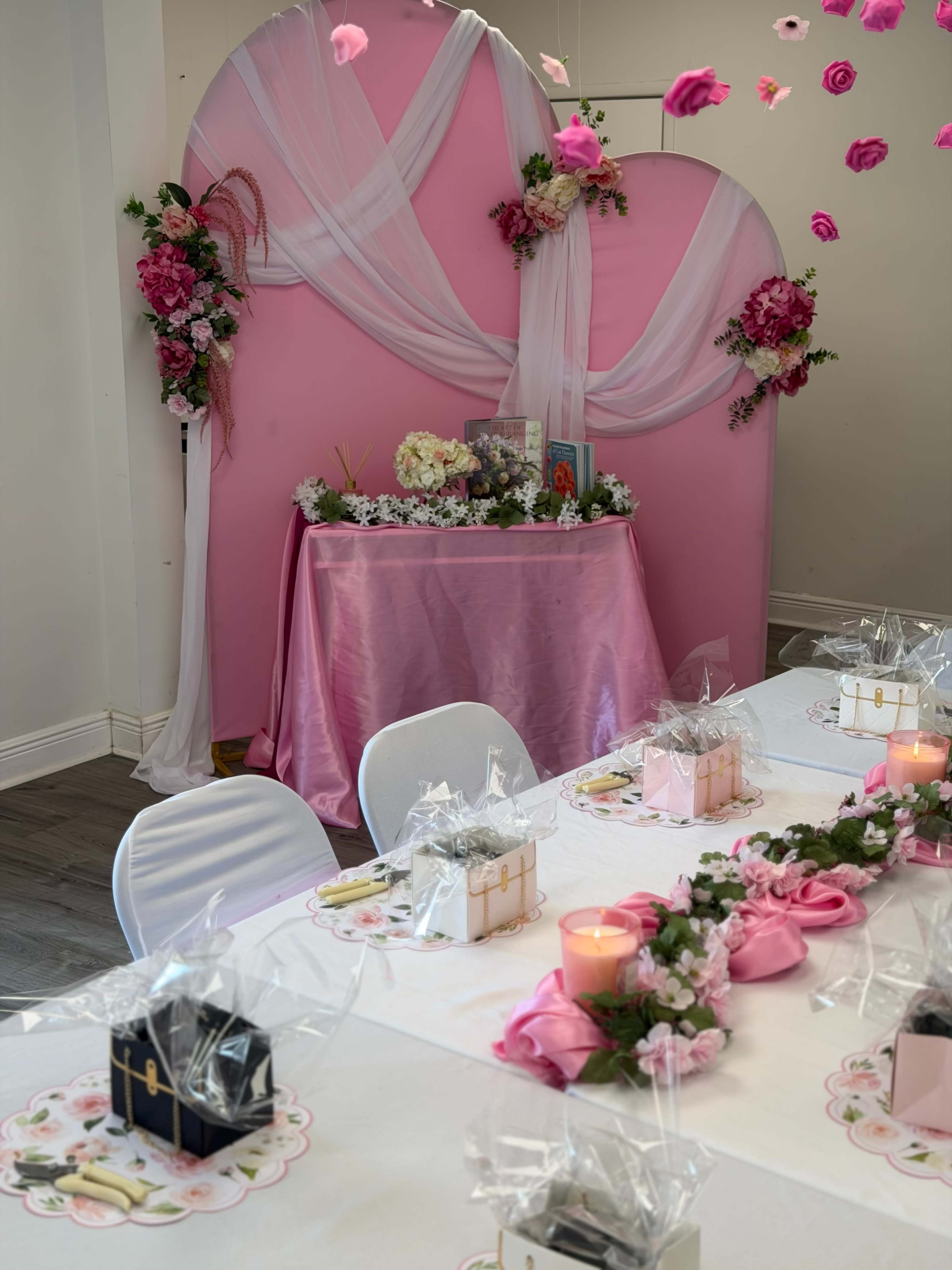 The image shows a decorated event space featuring a pink backdrop with floral arrangements, a table with elegant decorations, and a long dining table adorned with gifts and candles.