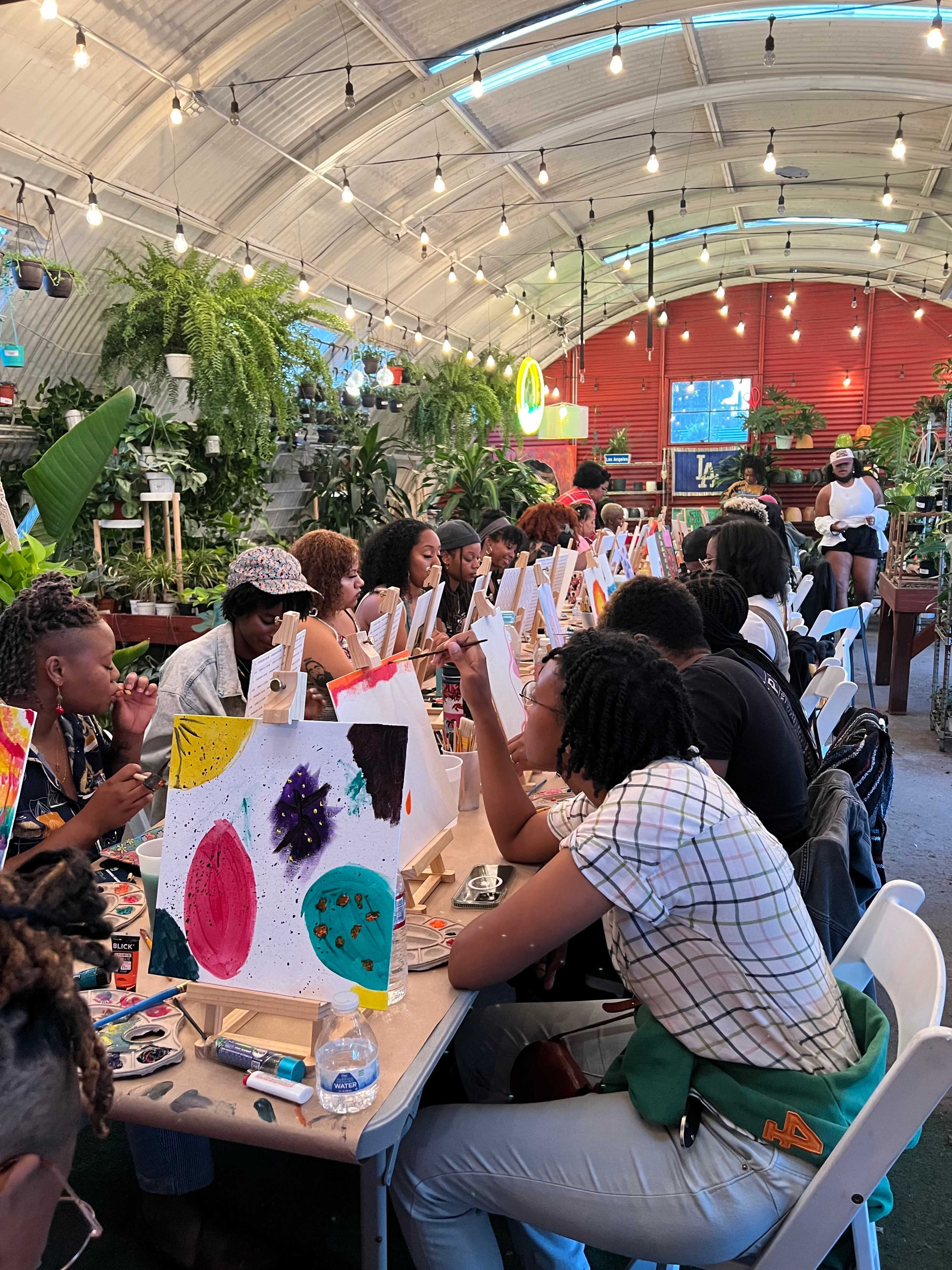 A group of people is painting on easels in a vibrant indoor space filled with plants and creative energy.