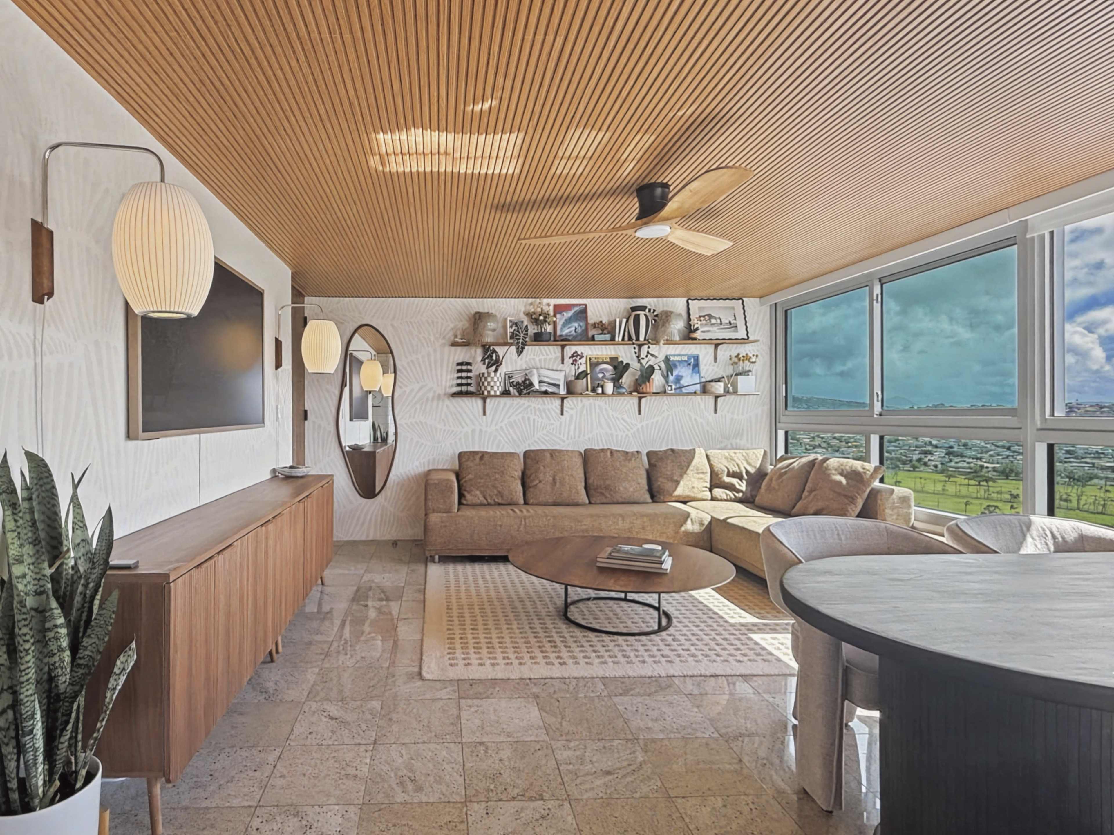 A modern living room features a sectional sofa, a large round coffee table, and a gallery wall with various framed pictures, all set against large windows offering a view of the outdoors.