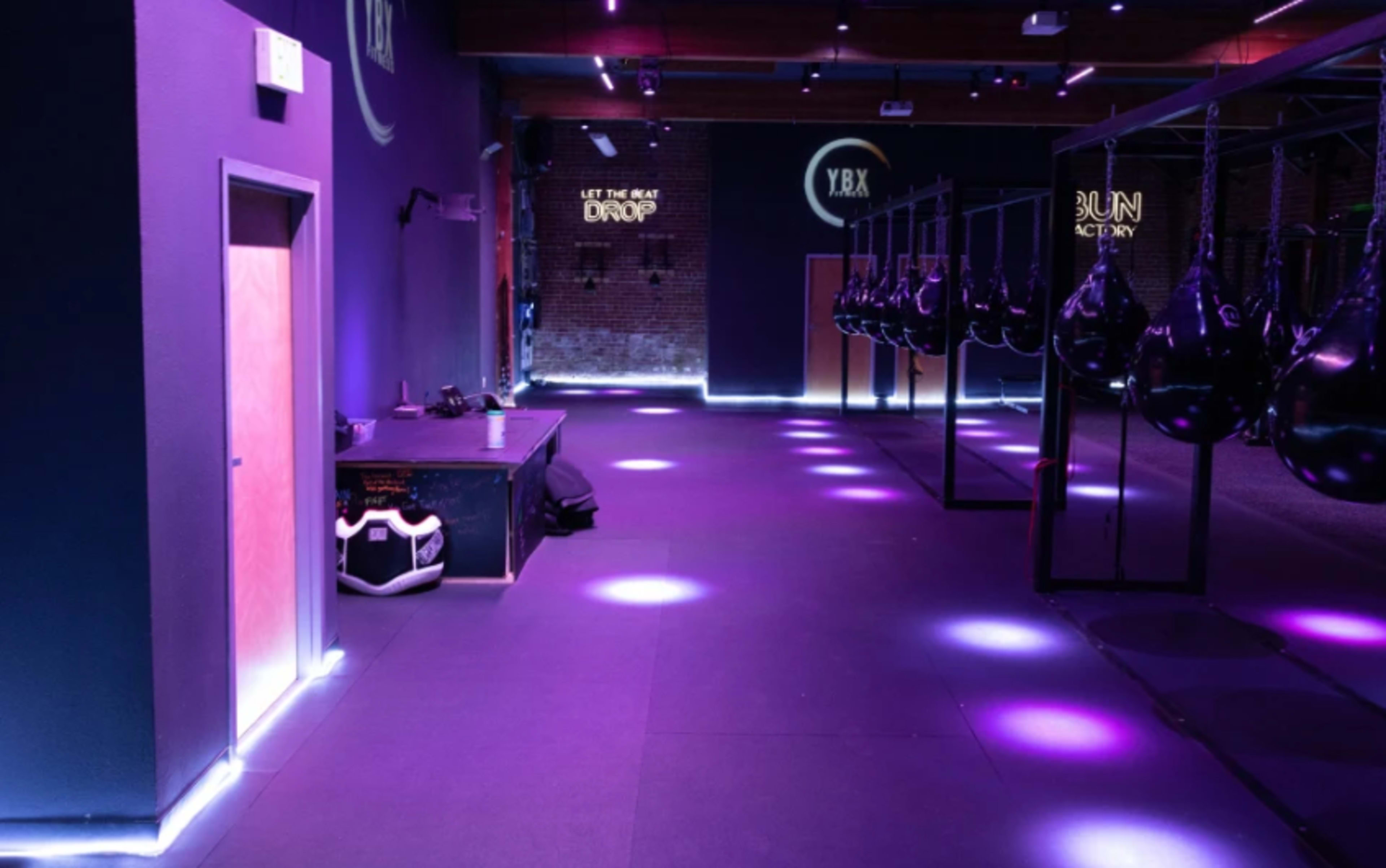 A dimly lit gym with purple lighting, featuring boxing bags suspended from a frame and a reception area in the corner.