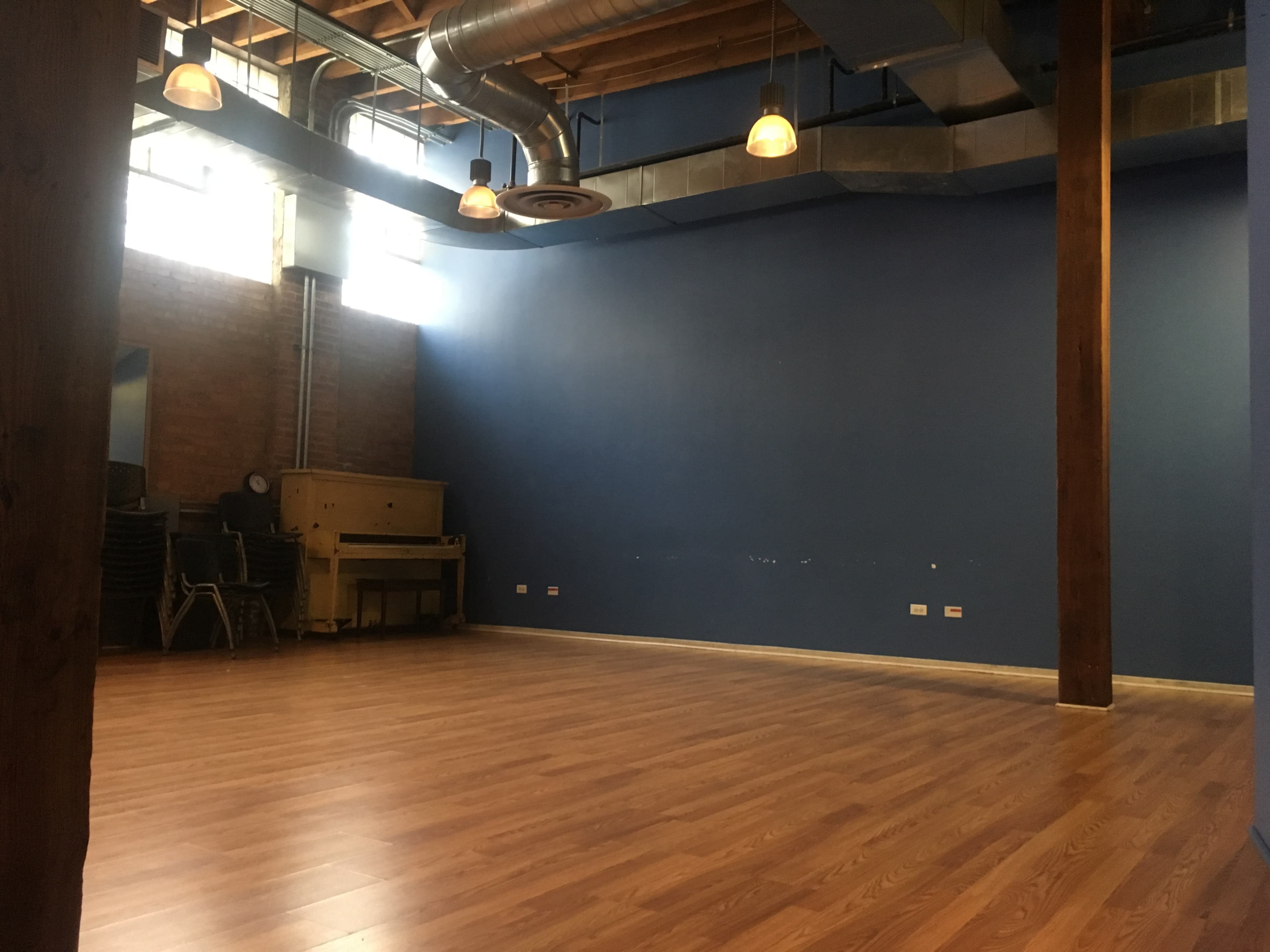 An empty room with wooden flooring, blue walls, and a row of stacked chairs against a wall.