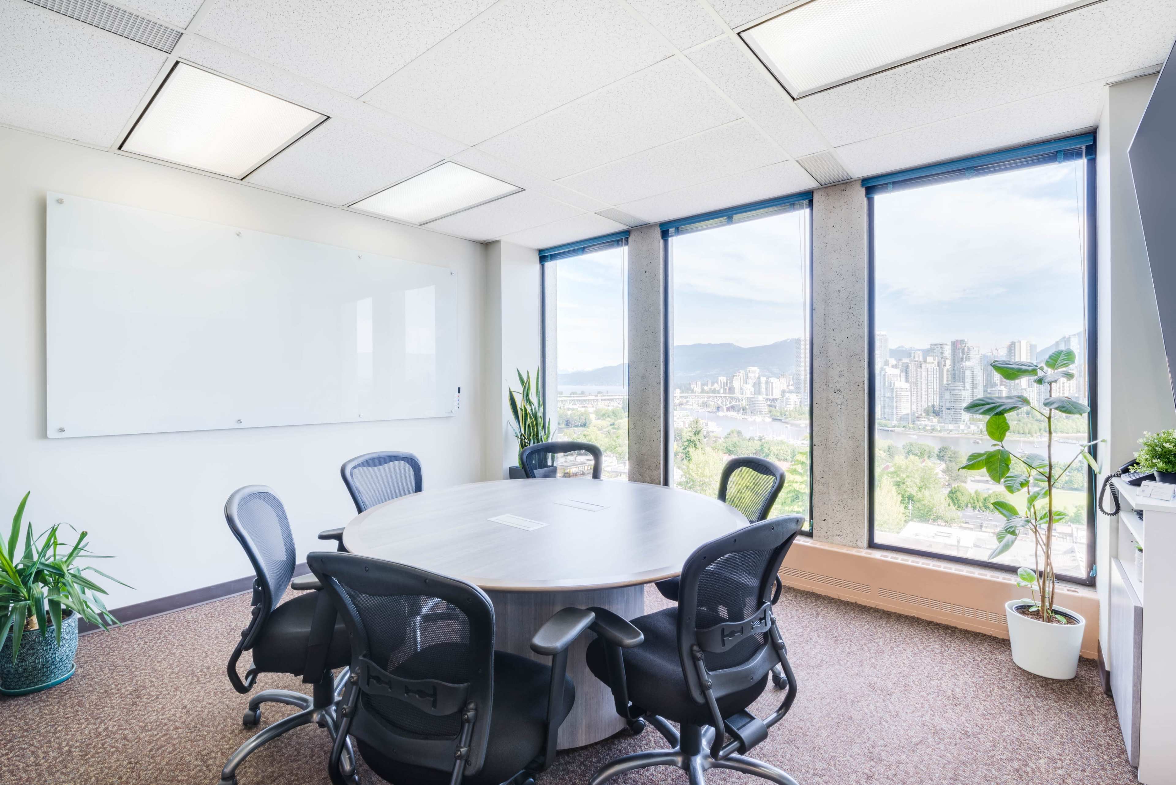 A modern conference room features a circular table surrounded by black chairs, large windows with city views, and plants in the corners.
