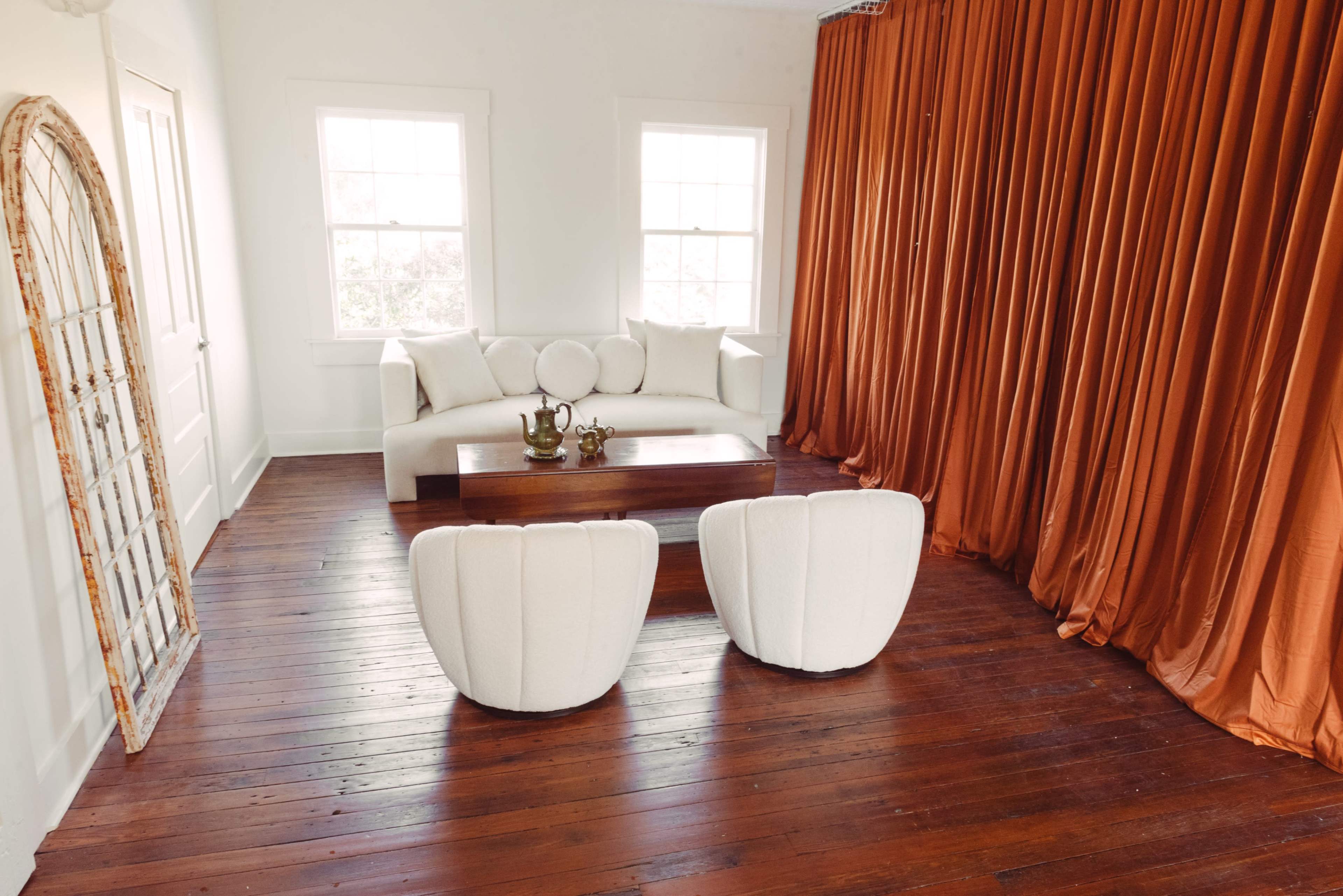 A bright room features a white sofa, two upholstered chairs, and a wooden coffee table, all set against warm, orange drapes and hardwood flooring.