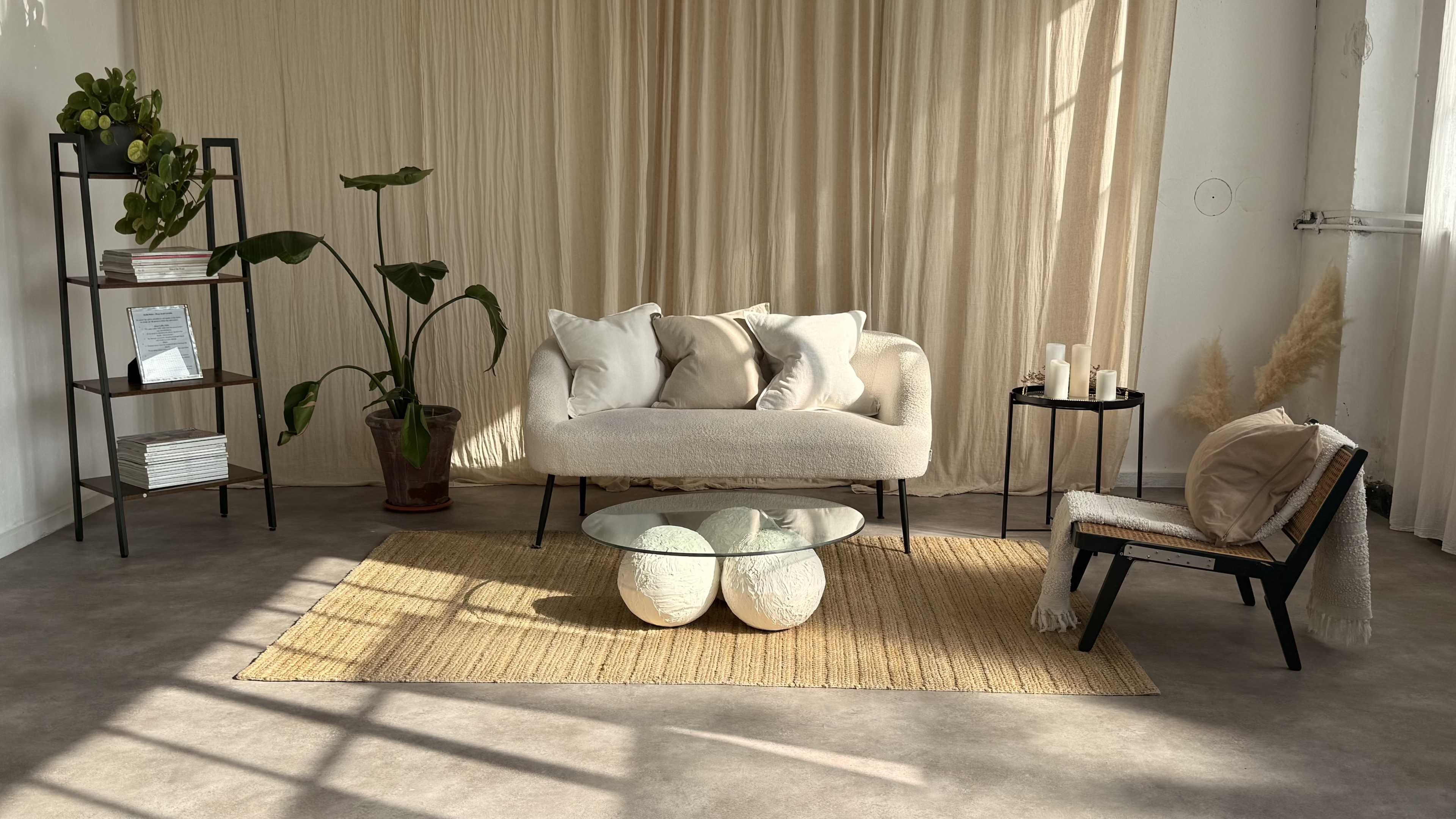 The image shows a minimalist living space featuring a light-colored sofa, a round glass coffee table, plants, and a small bookshelf, all set against a soft curtain backdrop.