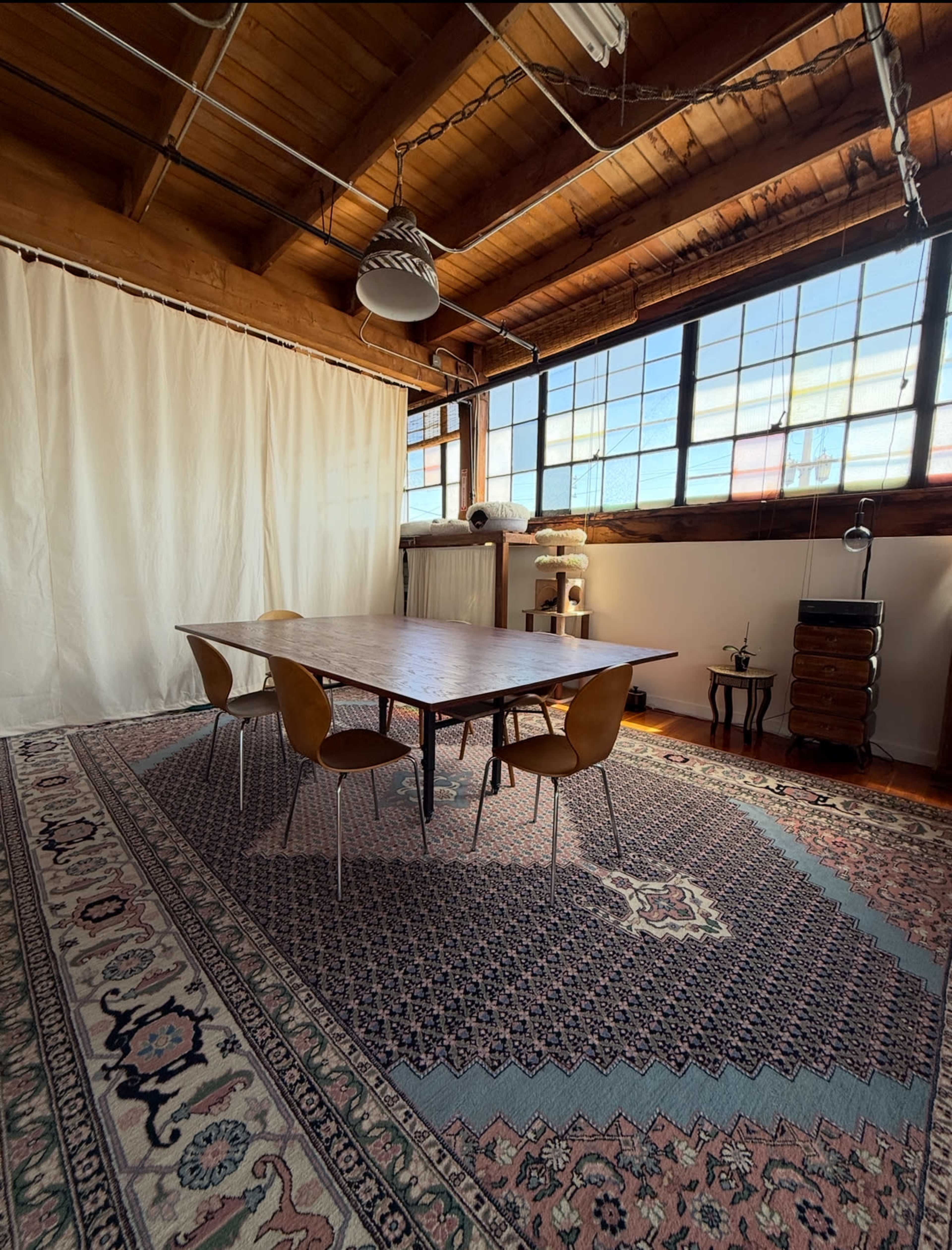 A spacious room features a wooden table surrounded by four brown chairs, with a large window letting in natural light, and an area rug covering the floor.