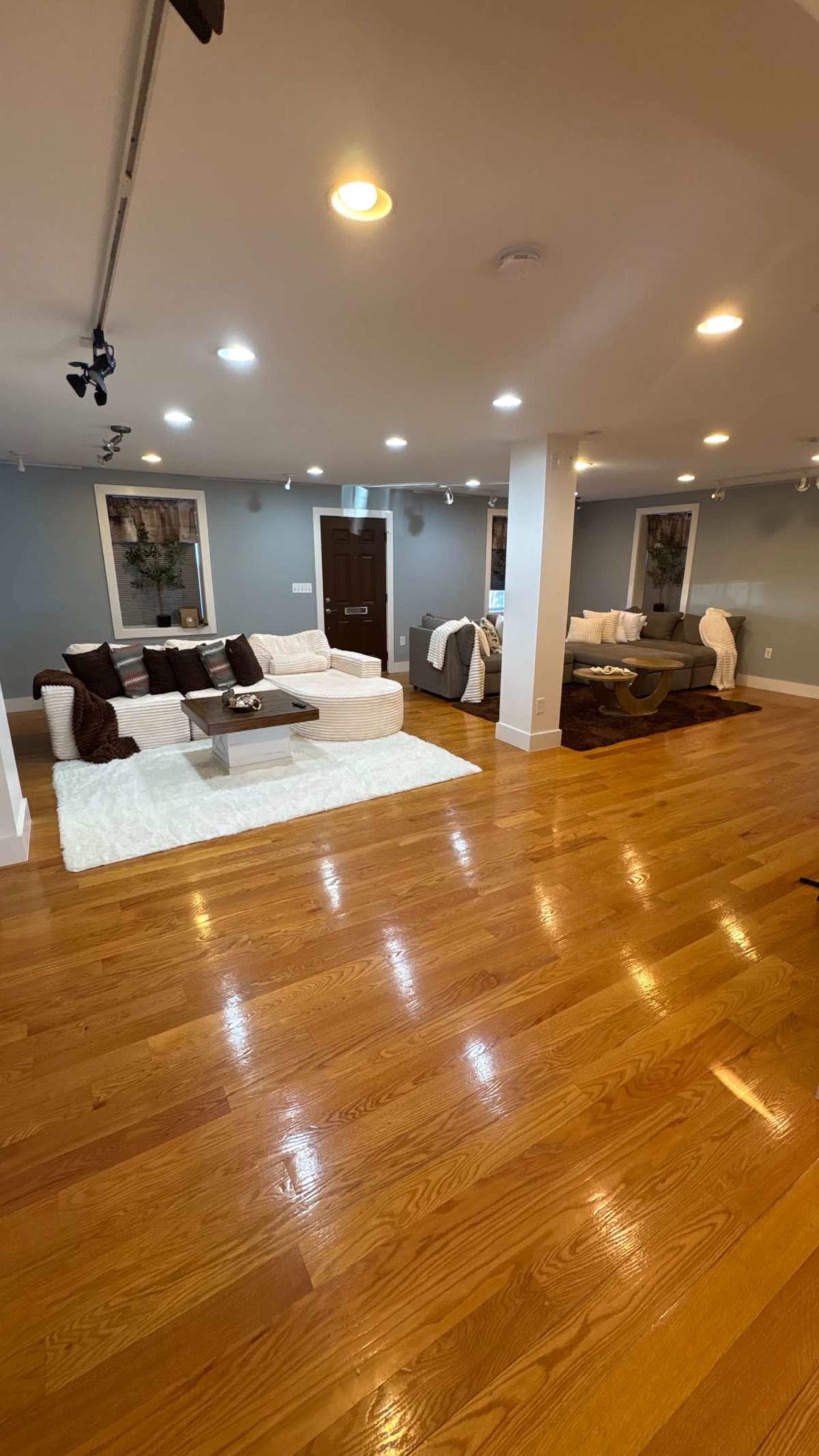 The image shows a spacious living room with hardwood floors, white couches, and a coffee table, illuminated by recessed lighting.