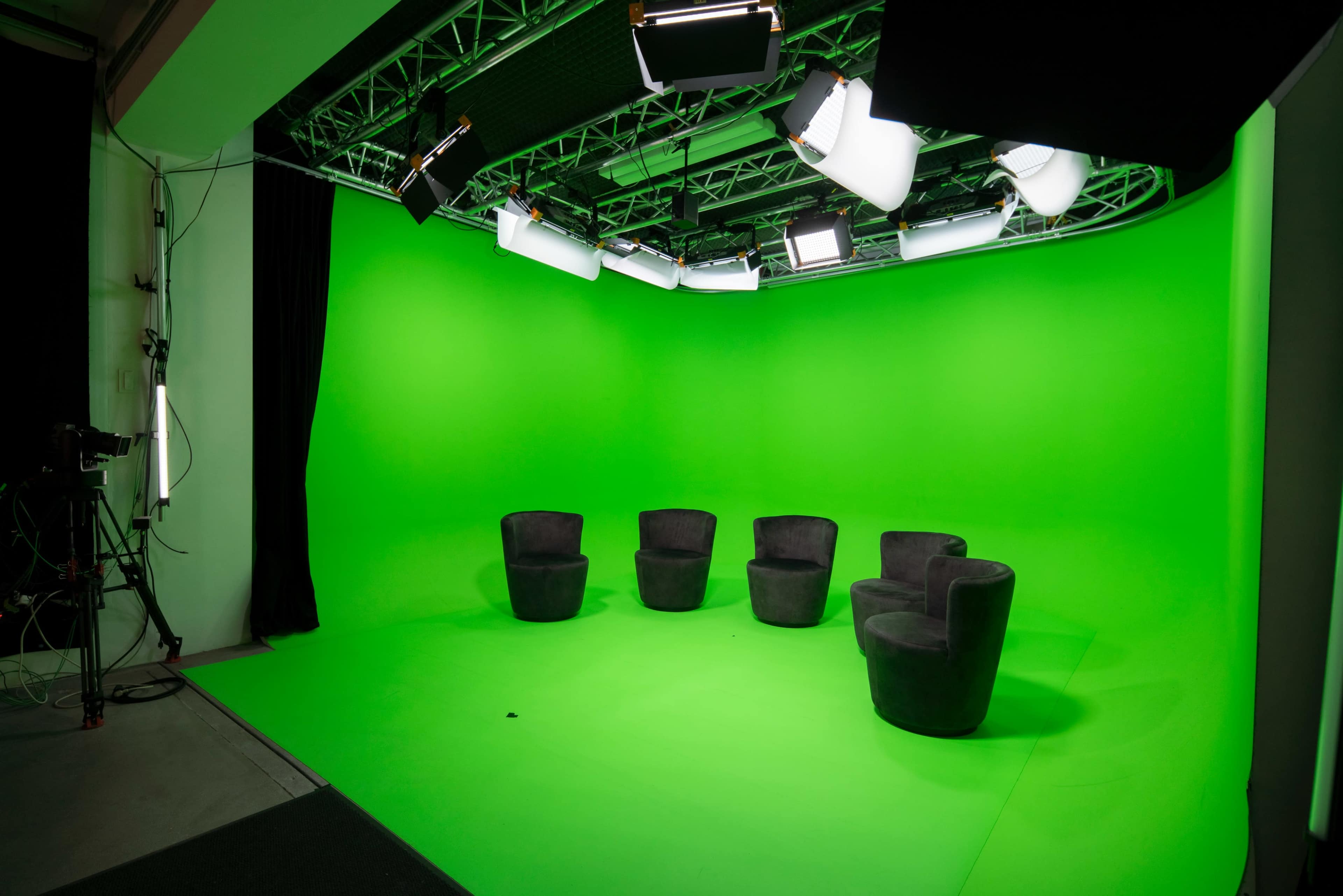 A studio setup with four black chairs arranged on a green screen backdrop and overhead lighting.