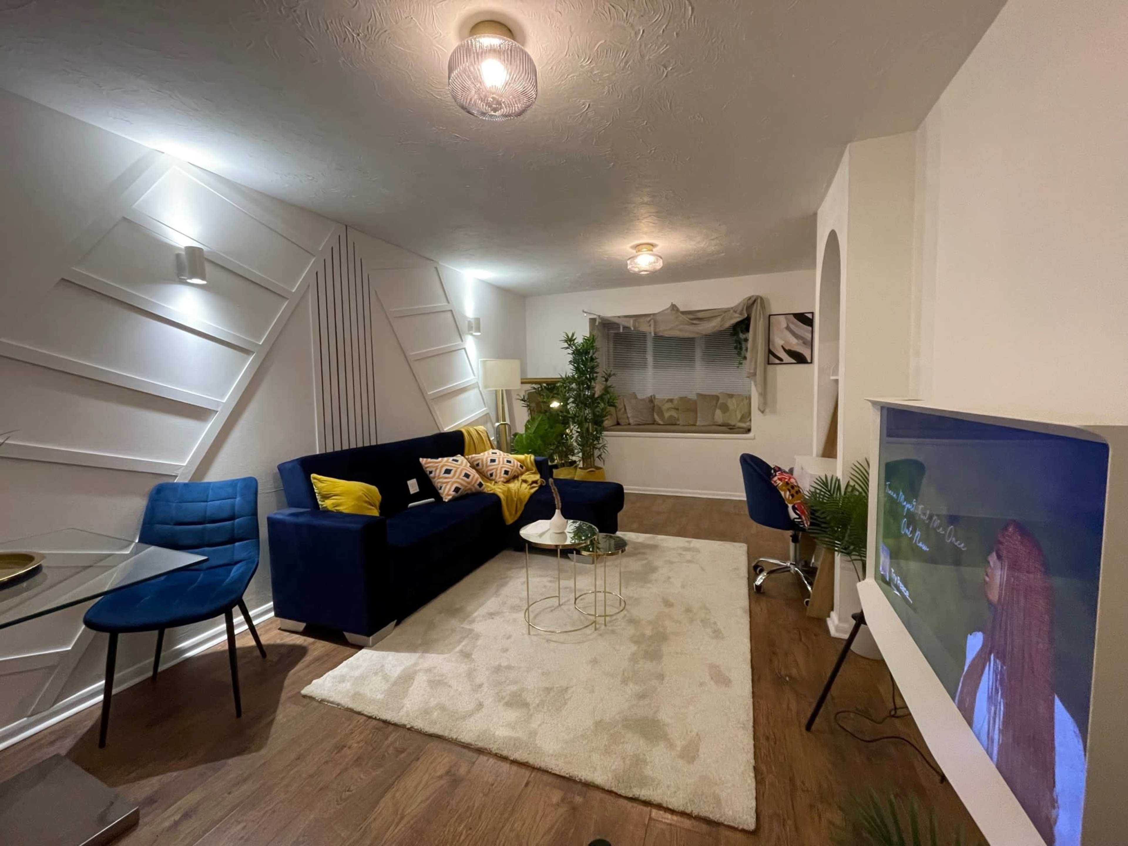The image shows a modern living room featuring a blue sofa, a glass table, and a television mounted on the wall, with a decorative rug on the floor and plants for accents.