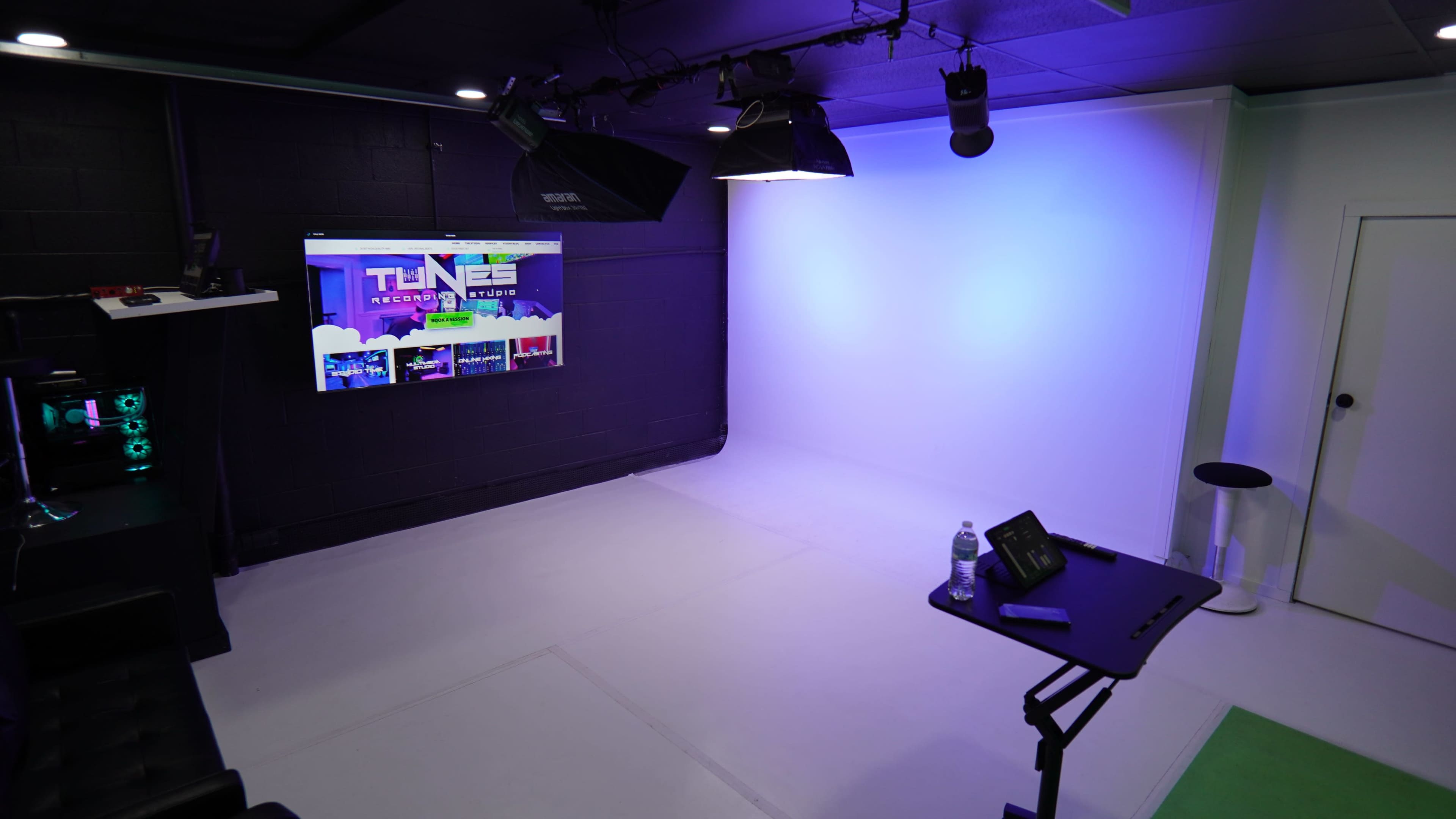 The image shows a recording studio with a blank white backdrop, a monitor displaying "TUNES RECORDING STUDIOS," and various lighting equipment.