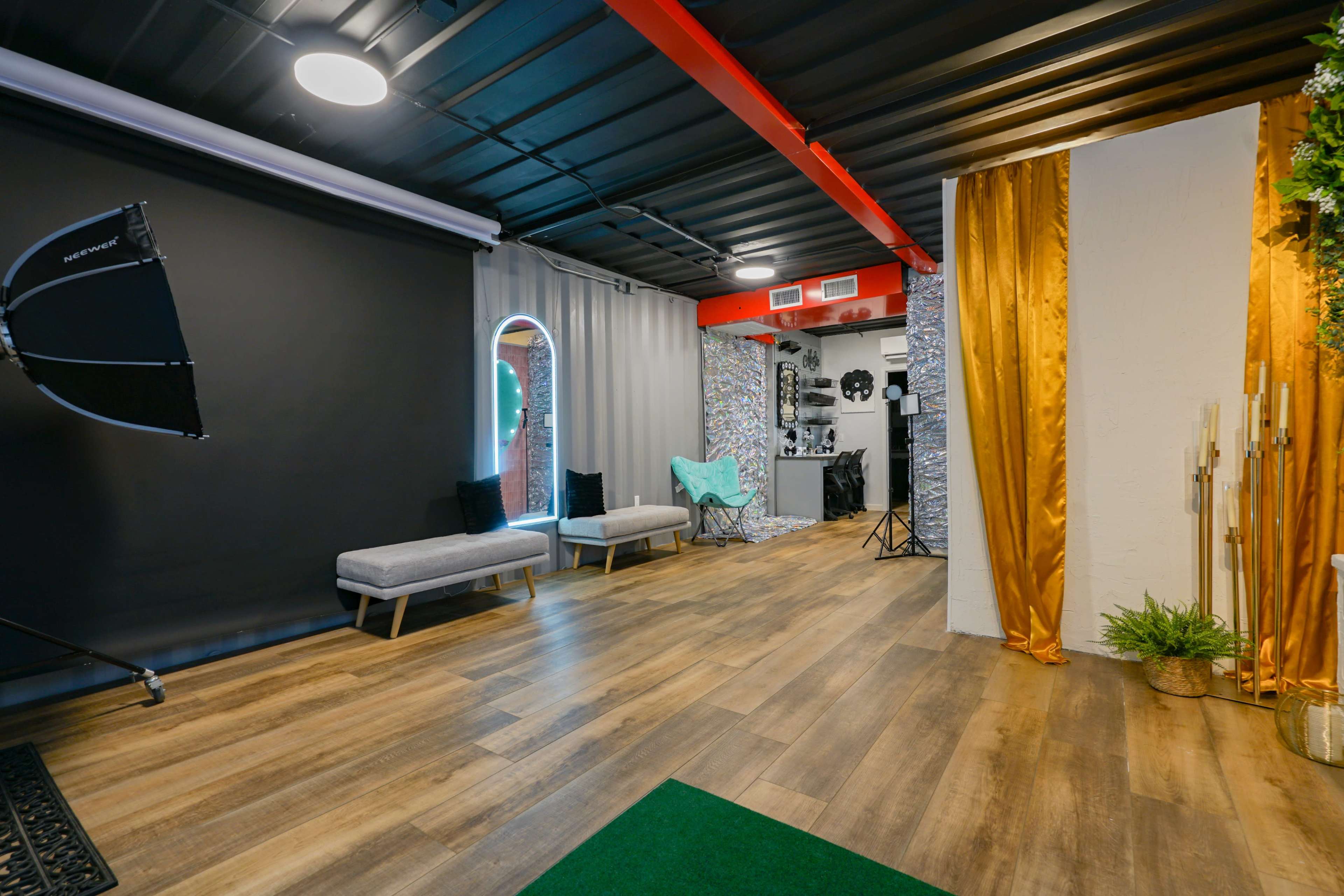 The image shows a modern studio space featuring a wooden floor, a black backdrop on one side, light-colored seating, and decorative elements including plants and drapes.