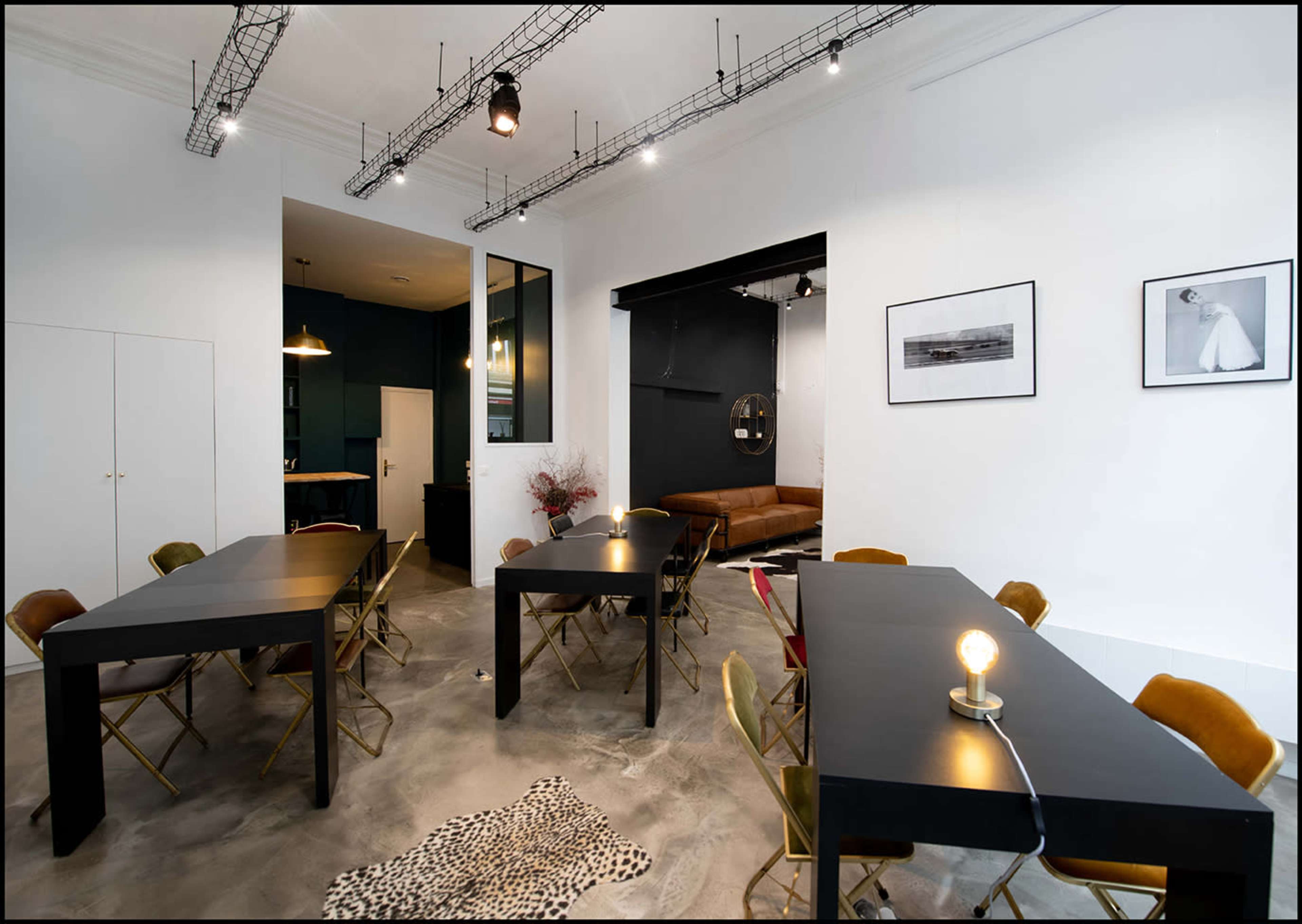 A modern workspace featuring several black tables with golden chairs, a sofa in the background, and minimalist decor on the walls.