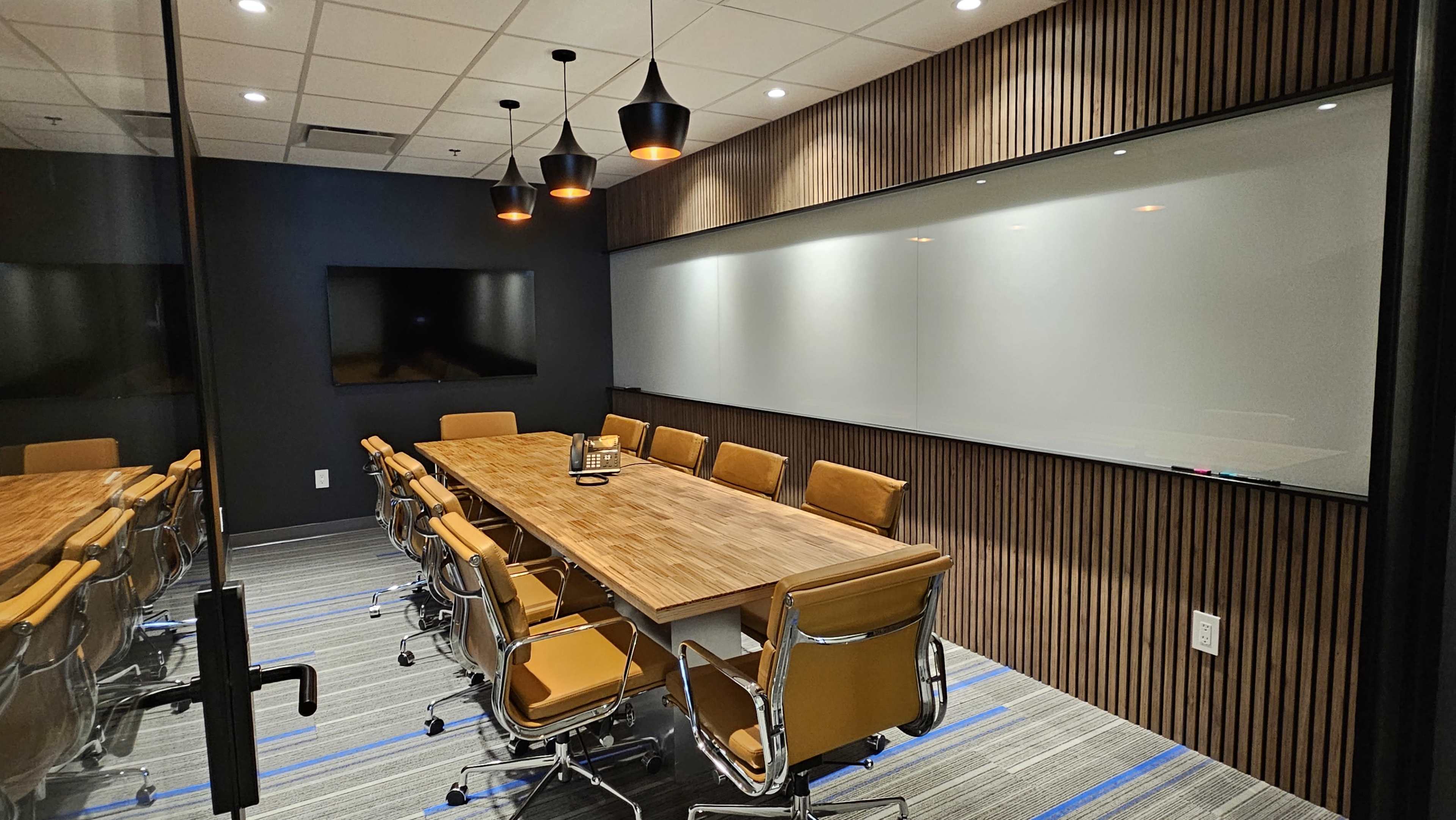The image shows a modern conference room with a long wooden table, orange leather chairs, a blank whiteboard, and a large screen on the wall.