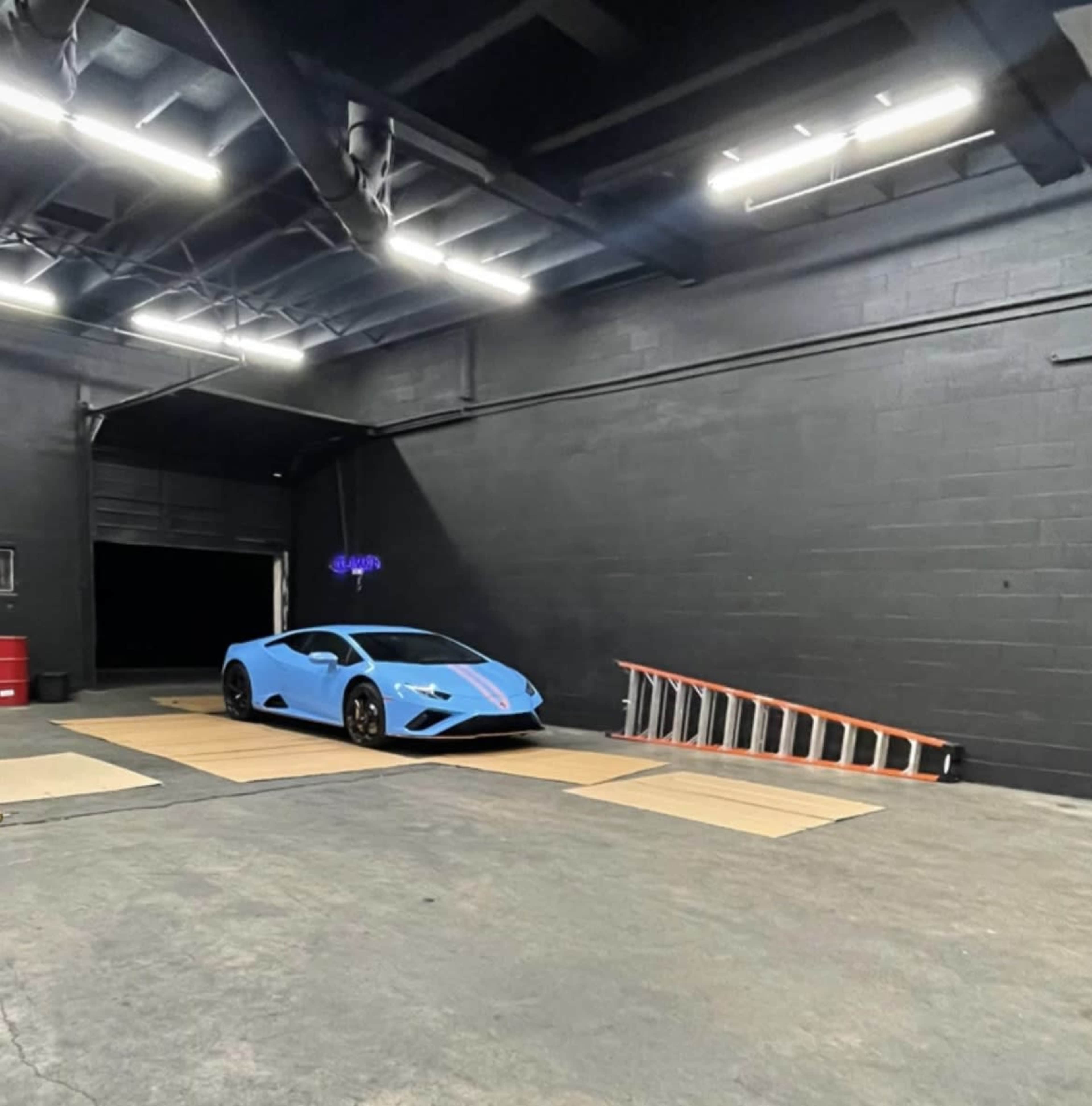 A blue sports car is parked in a dimly lit garage with a black wall and a wooden ramp nearby.