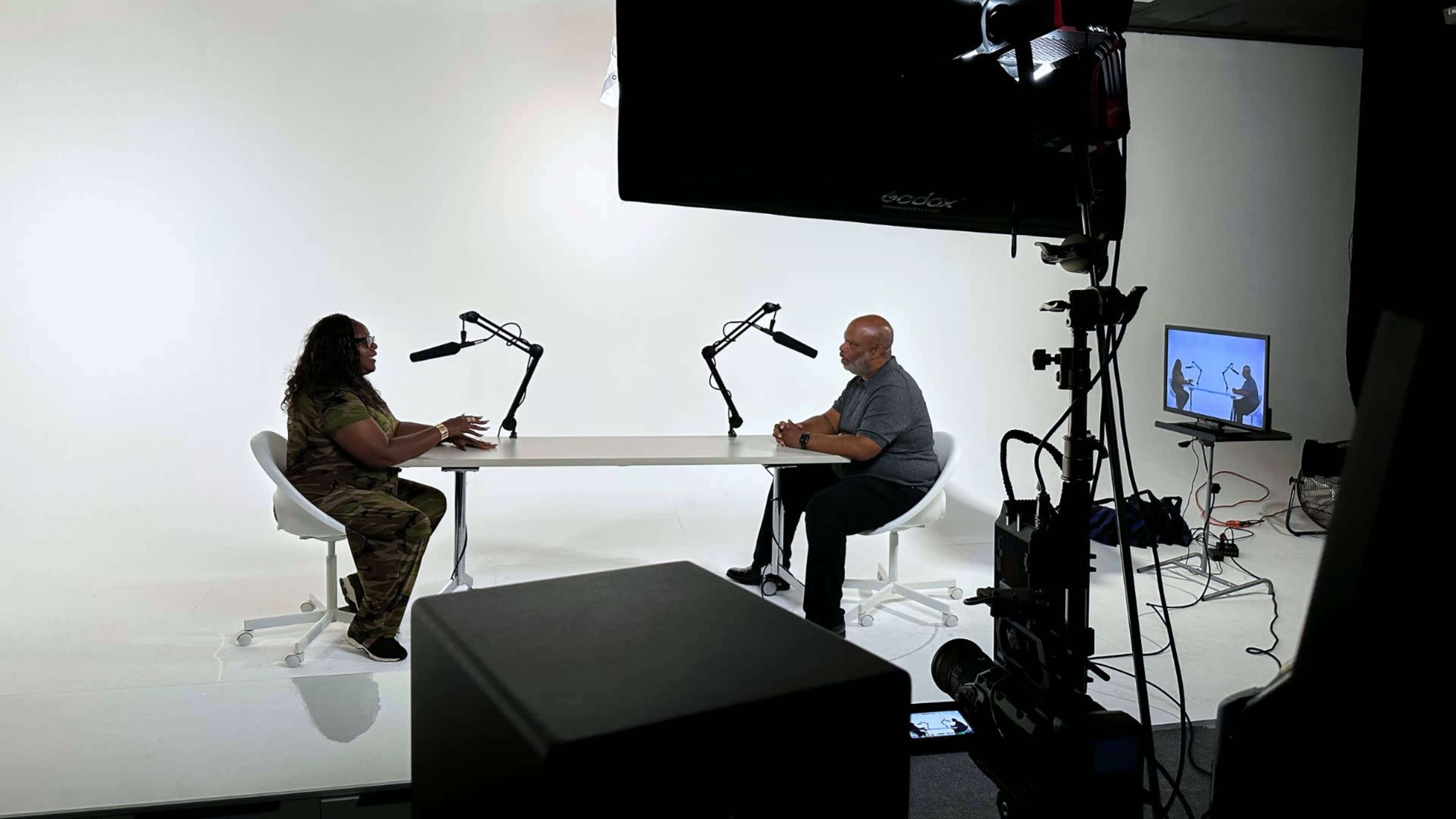 A woman in camouflage attire and a man in a gray shirt sit across from each other at a table in a minimalistic studio, with microphones positioned for a conversation.