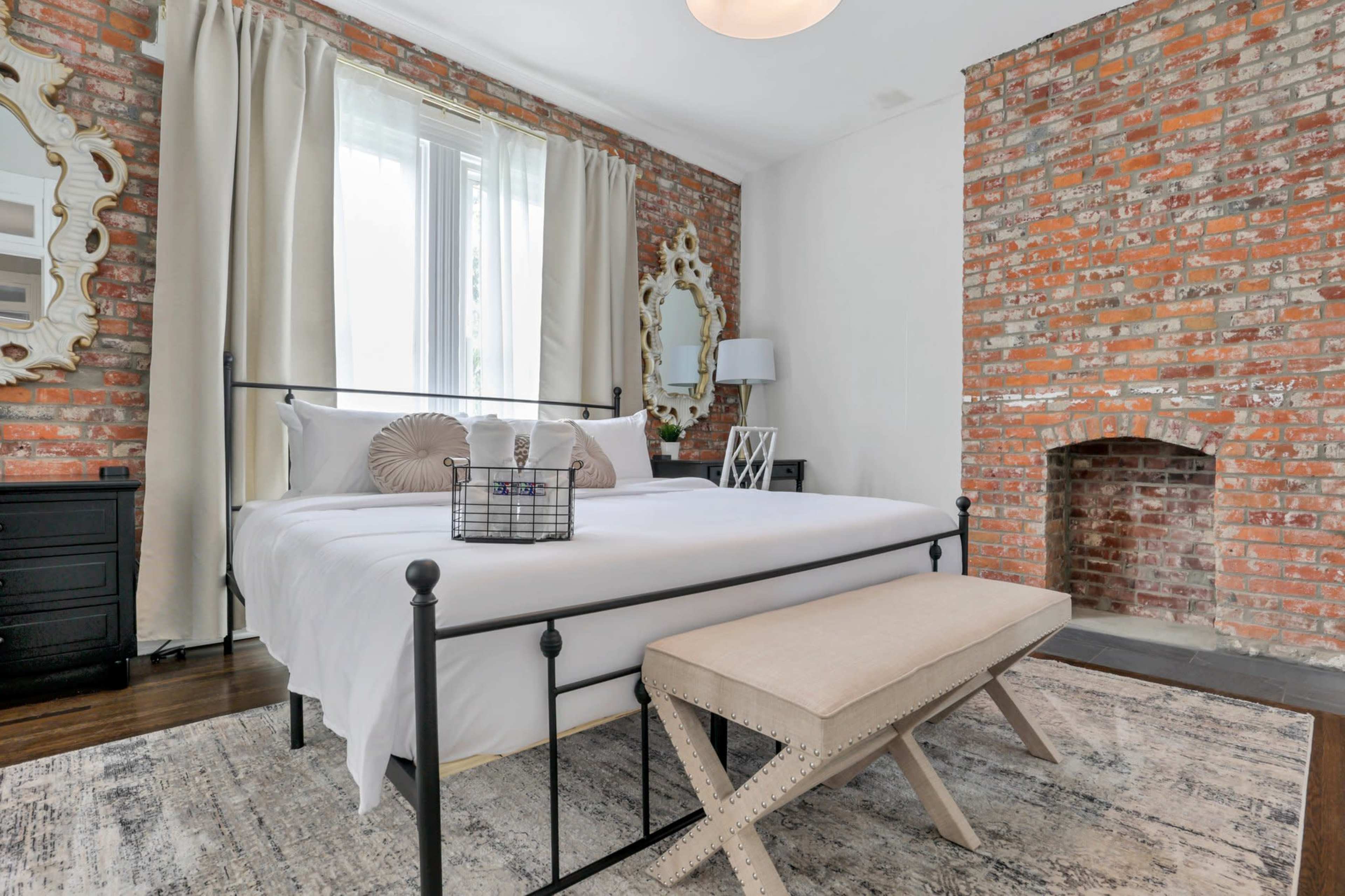 A bedroom featuring a metal-framed bed with white linens, a decorative bench at the foot, exposed brick walls, and large windows with sheer curtains.