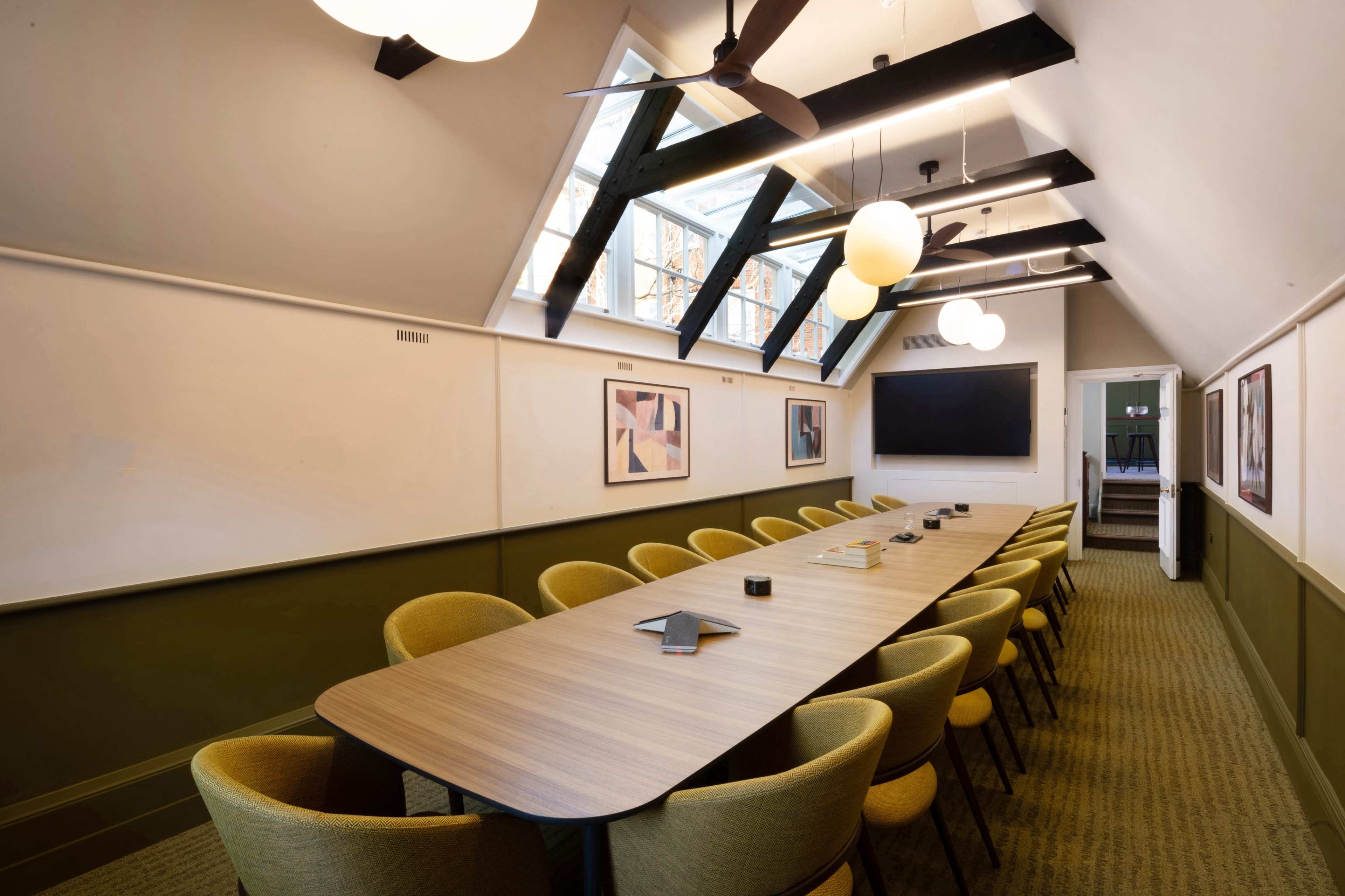 A long wooden conference table is surrounded by yellow chairs in a well-lit meeting room featuring large windows and a chalkboard.