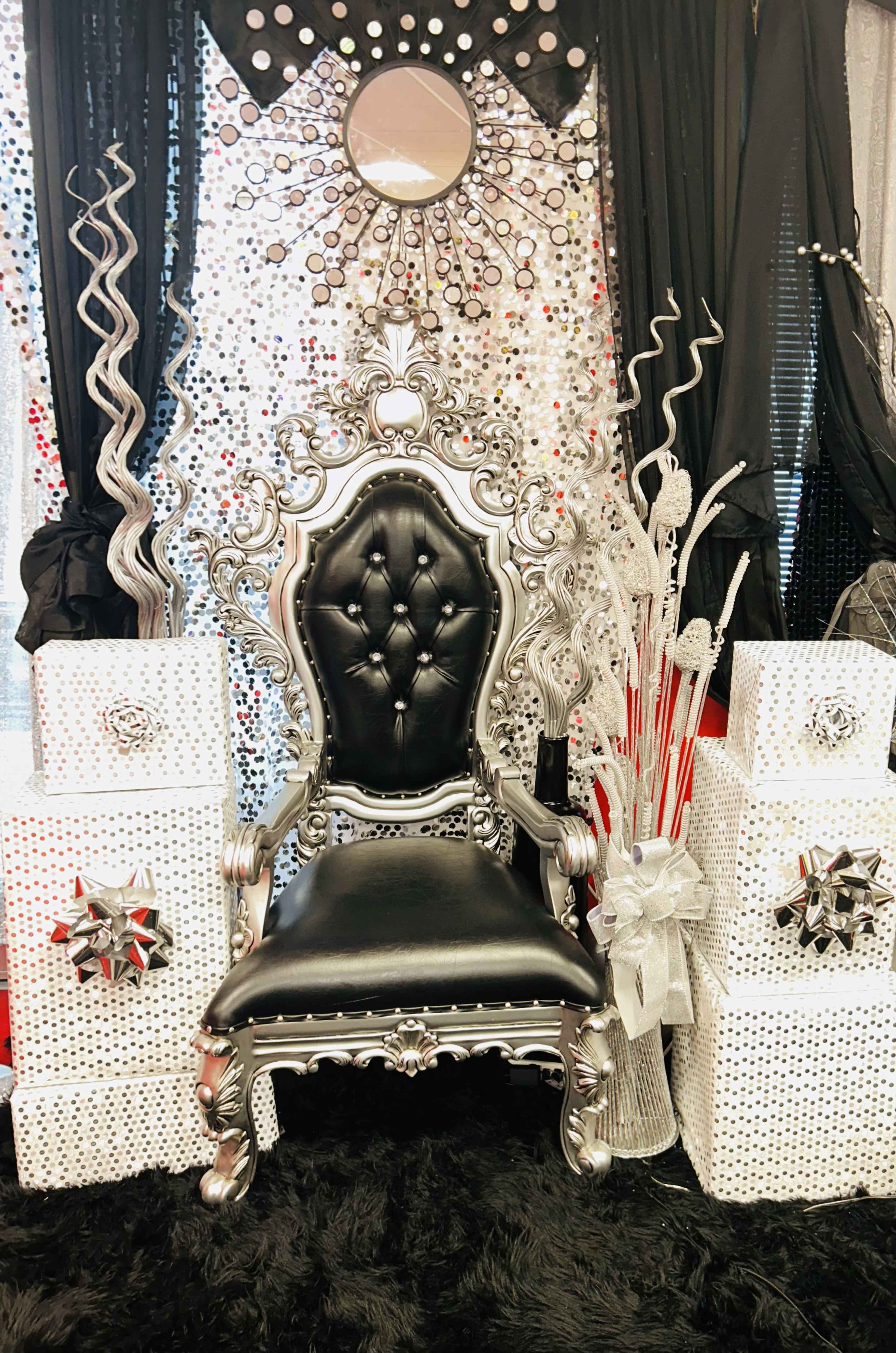 A large ornate silver chair with a black cushion is positioned against a backdrop of black curtains, sparkling sequins, and decorative white gift boxes.