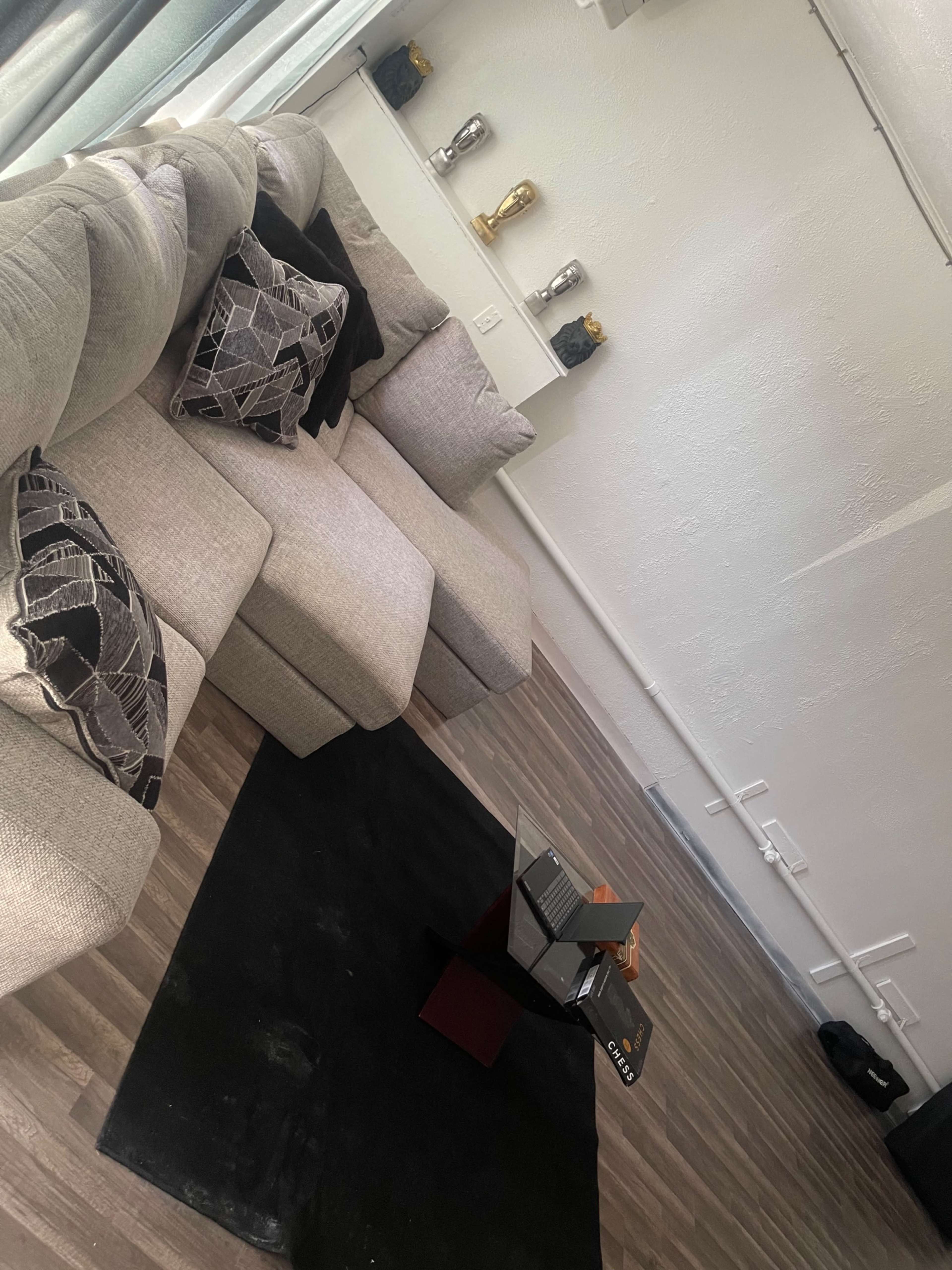 A minimalist living room with a beige sectional sofa, decorative cushions, a black rug, and a low coffee table, all against a light-colored wall.