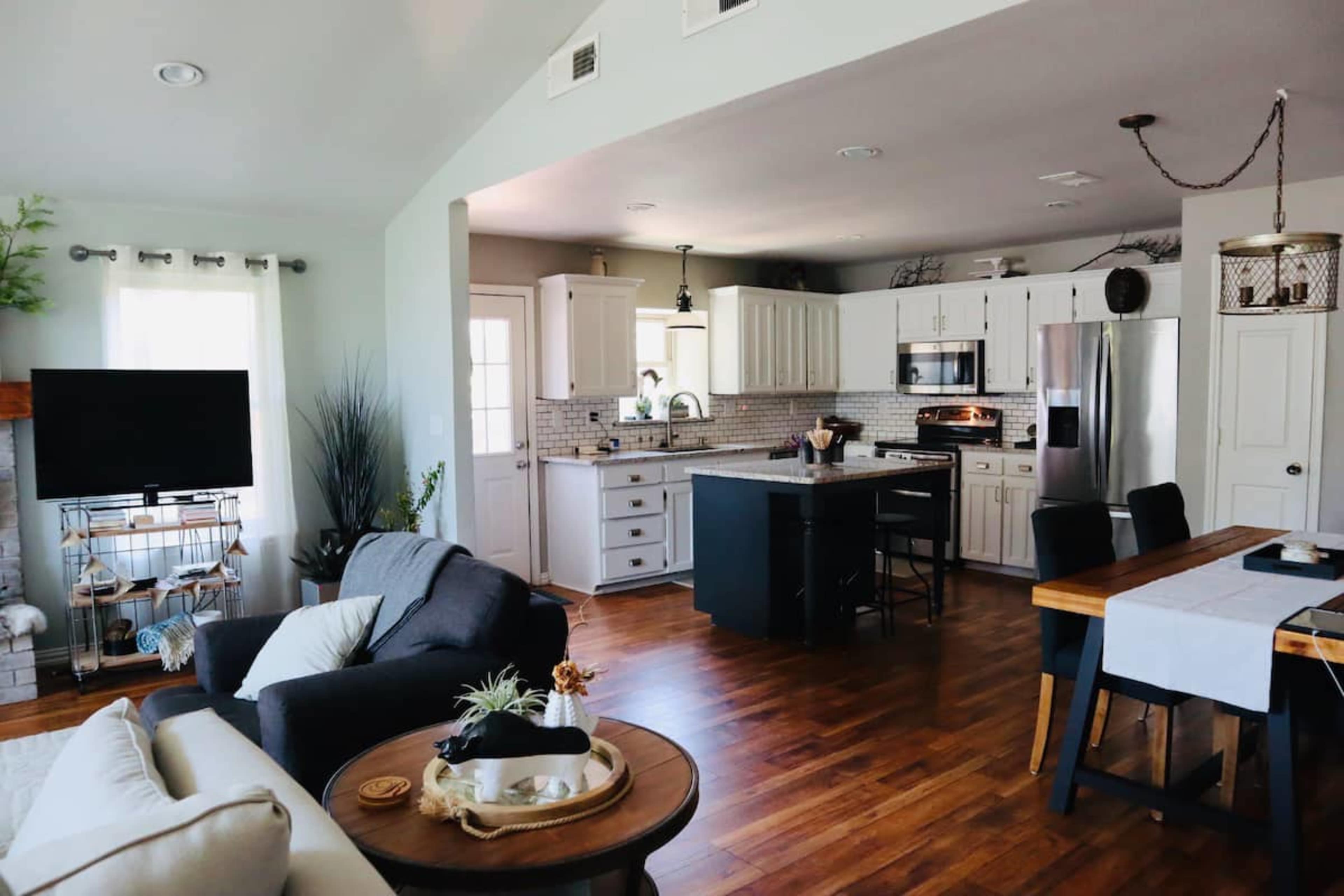A modern kitchen and living area with wooden flooring, white cabinetry, a gray island, and a dining table set for meals.