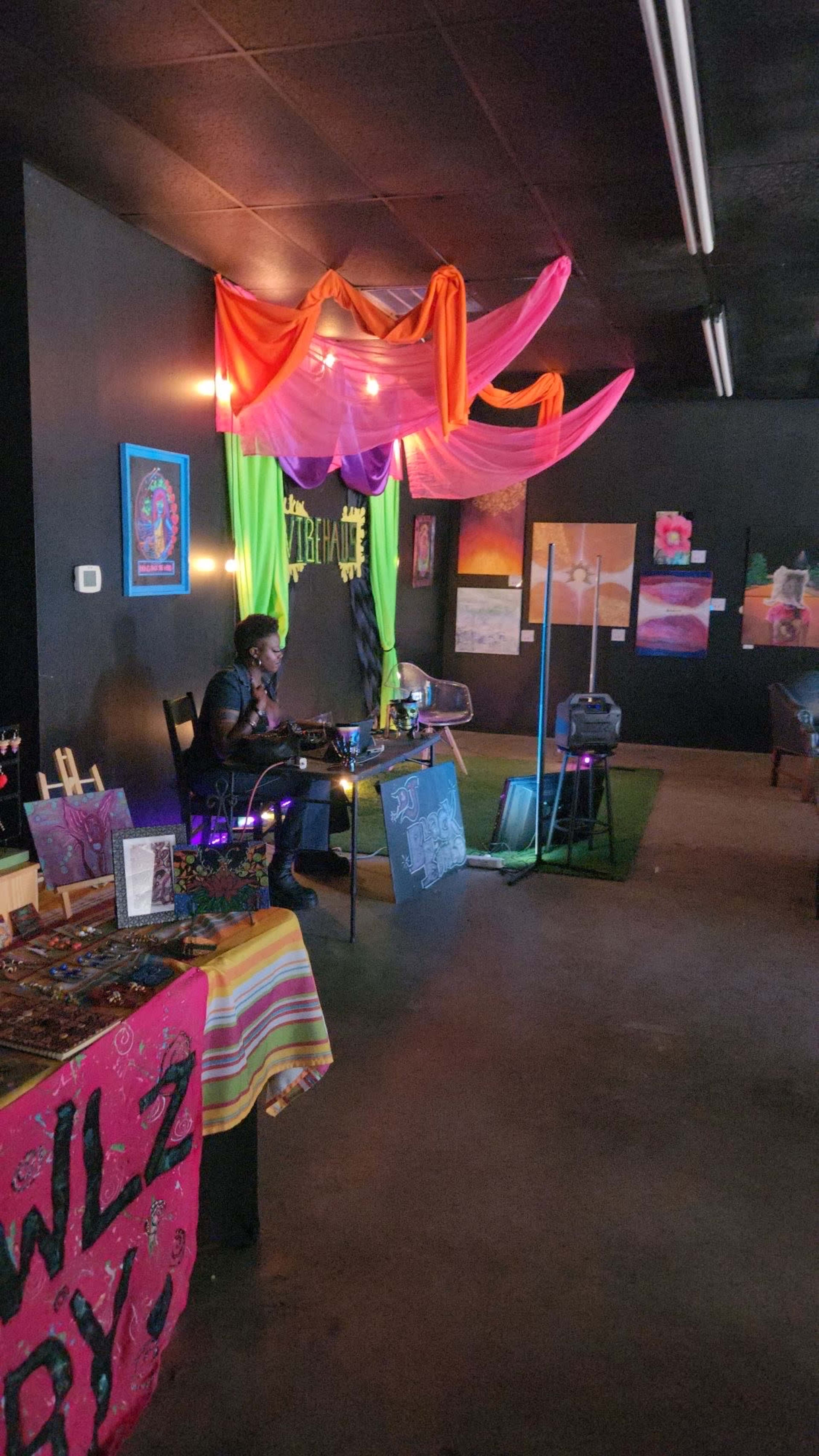 A colorful artistic space features a vendor's table on the left and vibrant fabric drapery overhead, with artworks displayed on the walls.