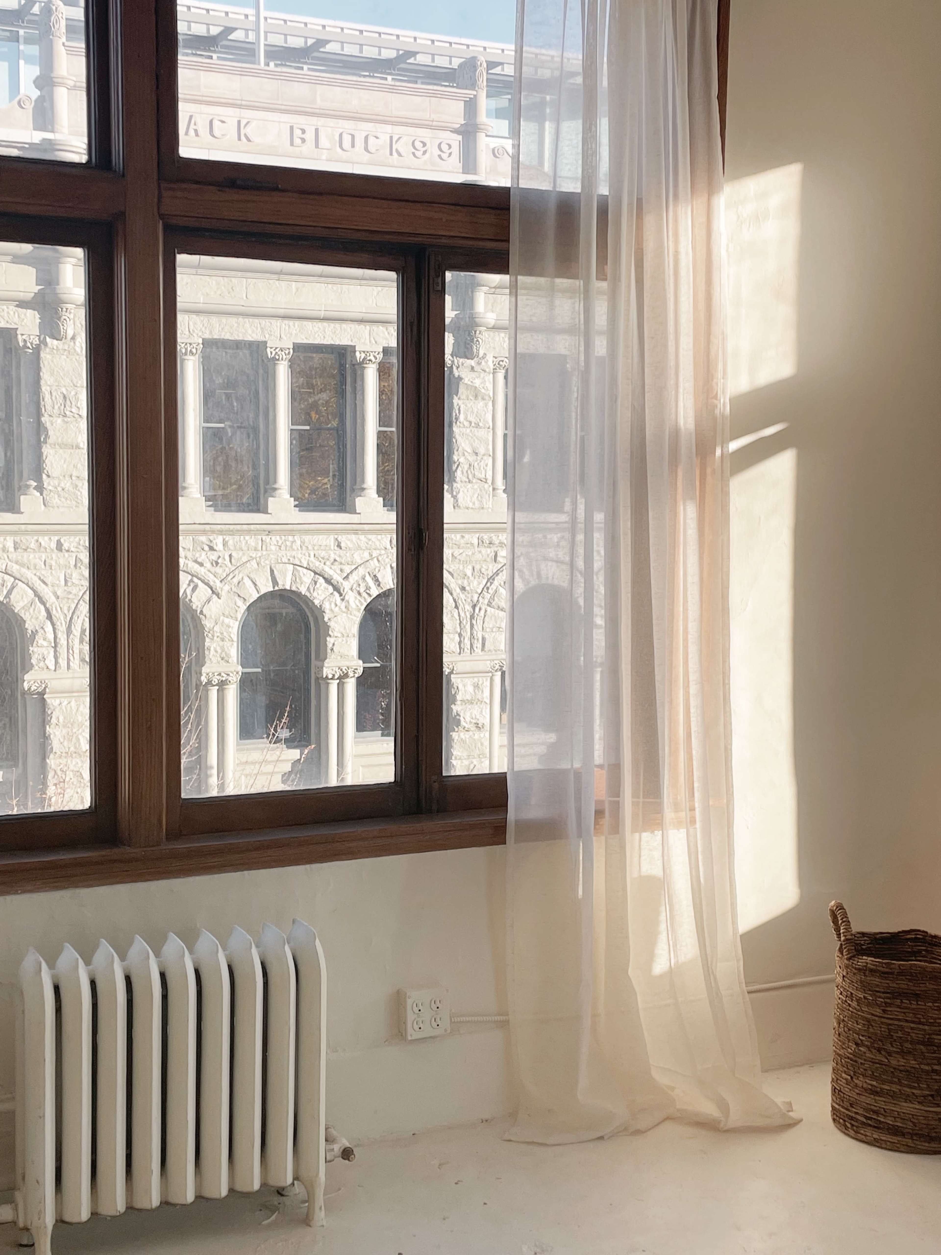 Light streams through a large window with sheer curtains, illuminating a room with a radiator and a woven basket, while revealing an architectural stone building outside.