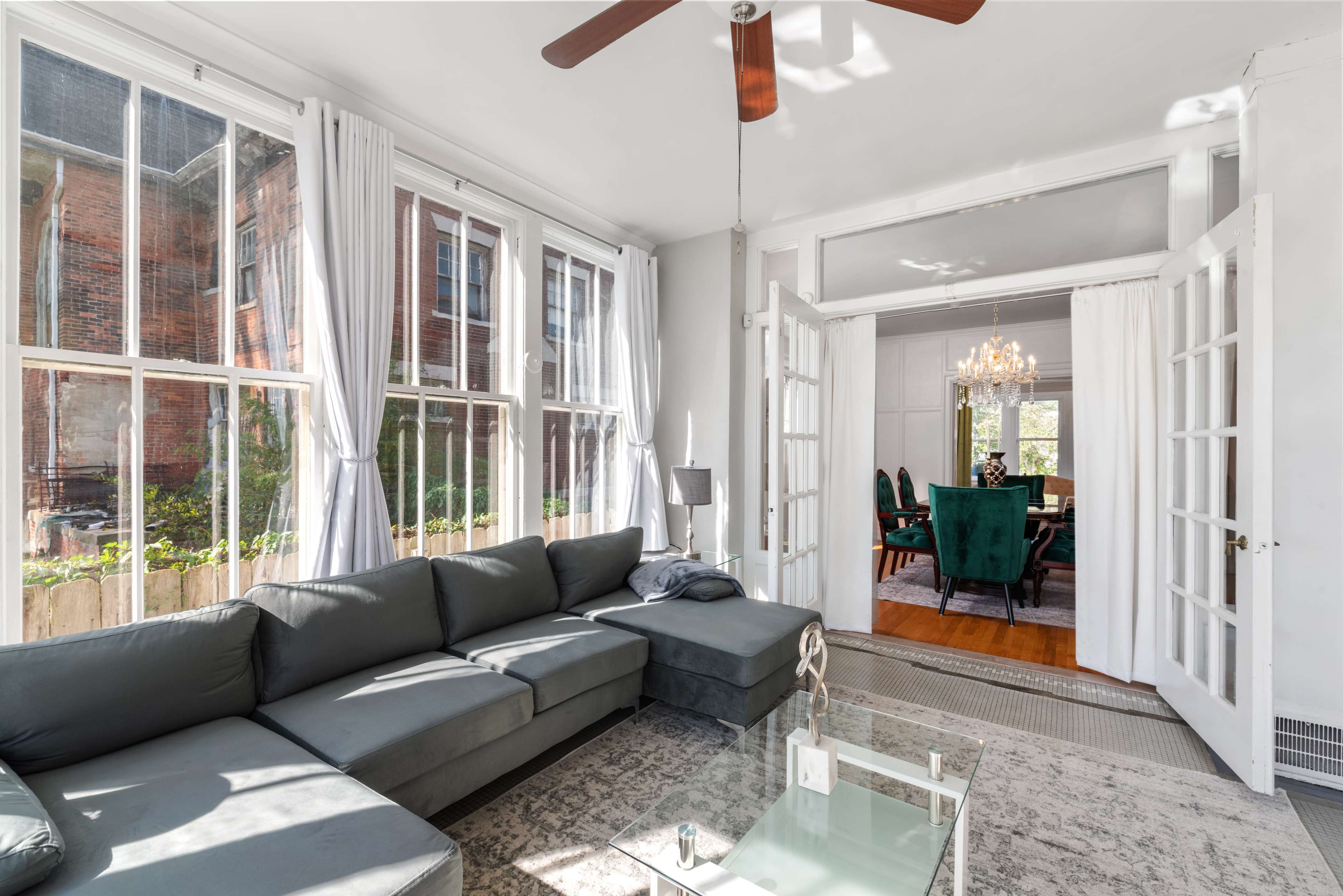 A modern living room with large windows, a gray sectional sofa, and open French doors leading to a dining area with green chairs.