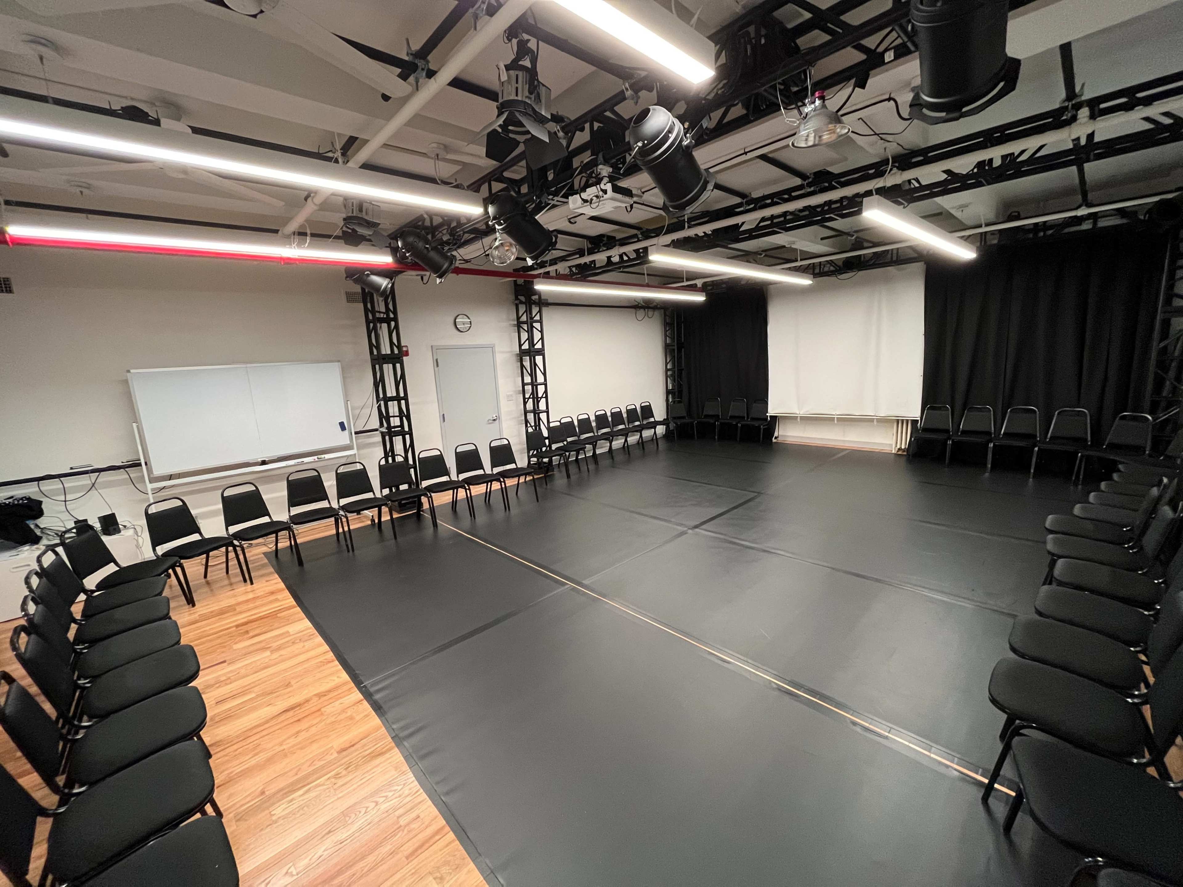 A rehearsal space with a black floor mat, surrounded by rows of black chairs arranged in a circular pattern against white and black walls.