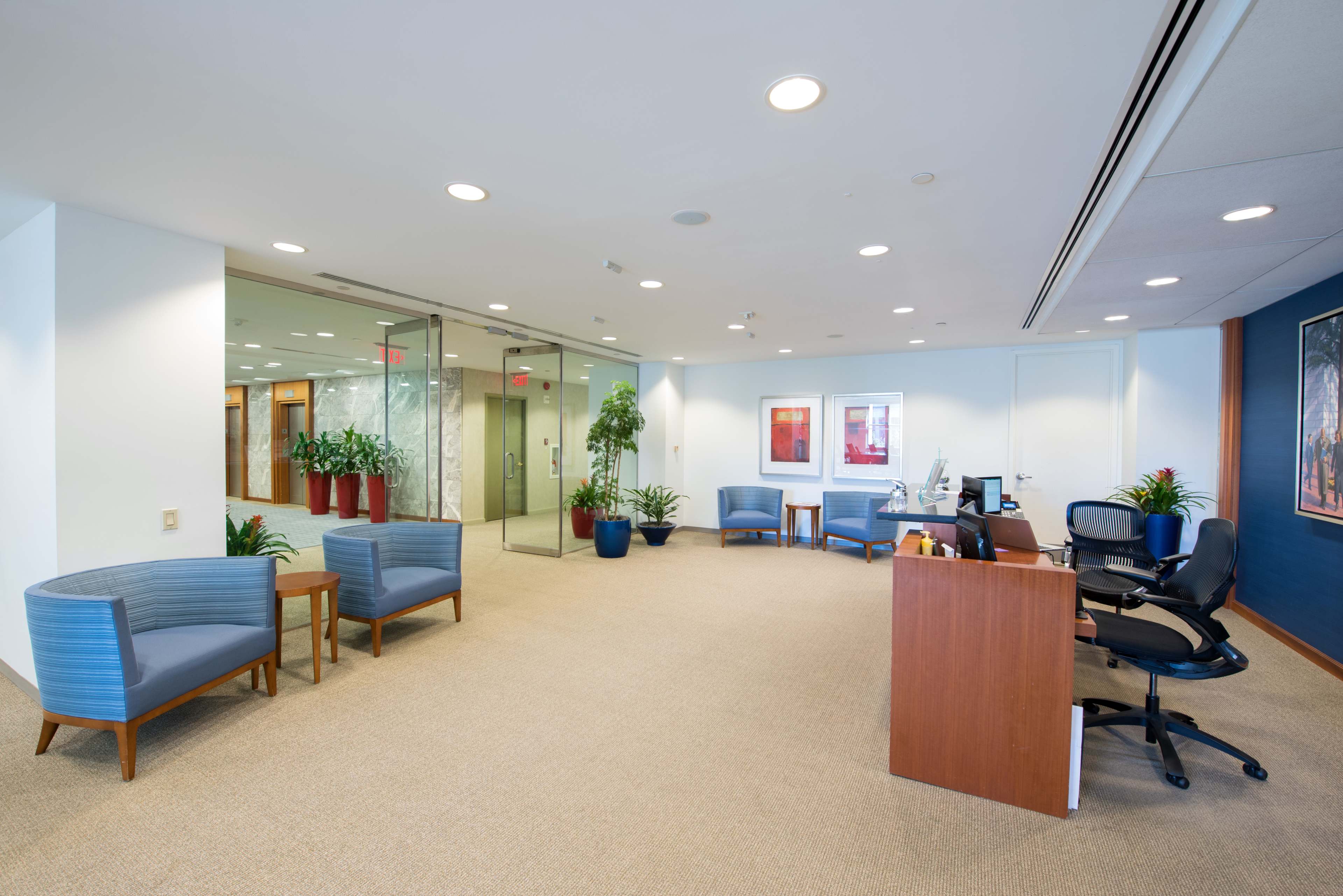 A modern office reception area with blue seating, potted plants, and a front desk facing a glass-walled meeting room.