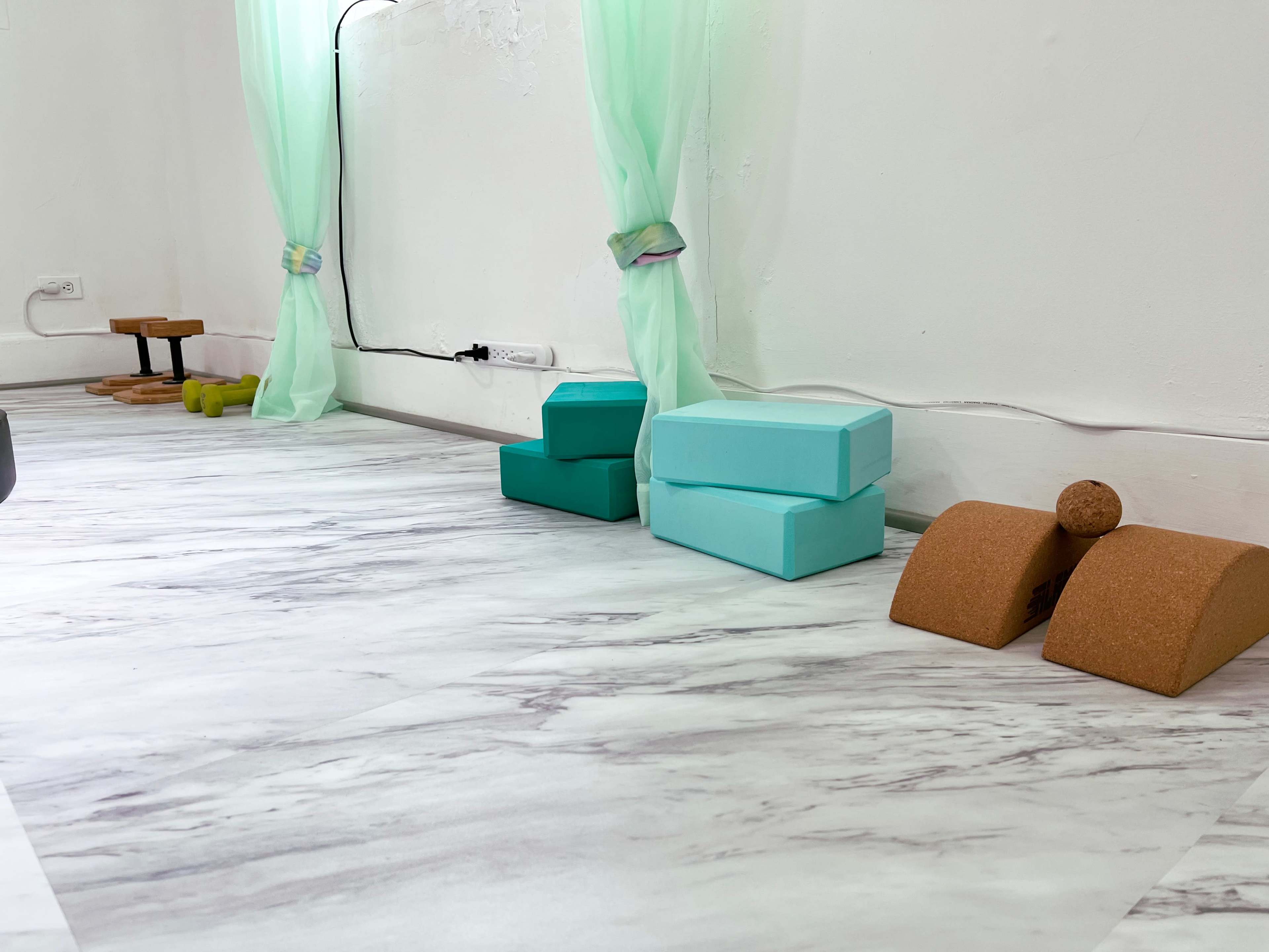 A light-colored marble floor with several yoga props, including turquoise blocks and cork accessories, near a wall adorned with sheer curtains.