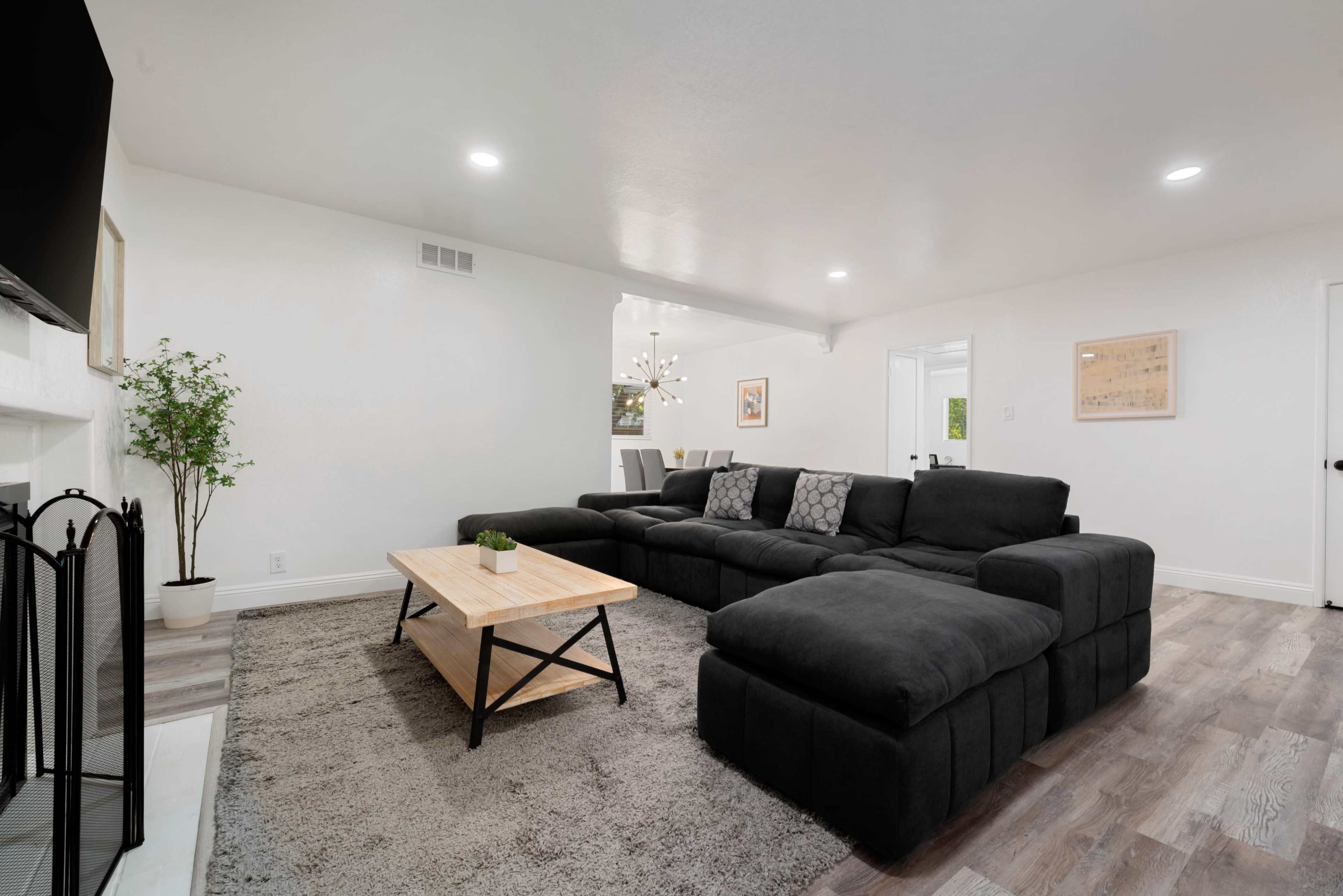 A modern living room with a large black sectional sofa, a wooden coffee table, and a plush area rug, illuminated by ceiling lights.