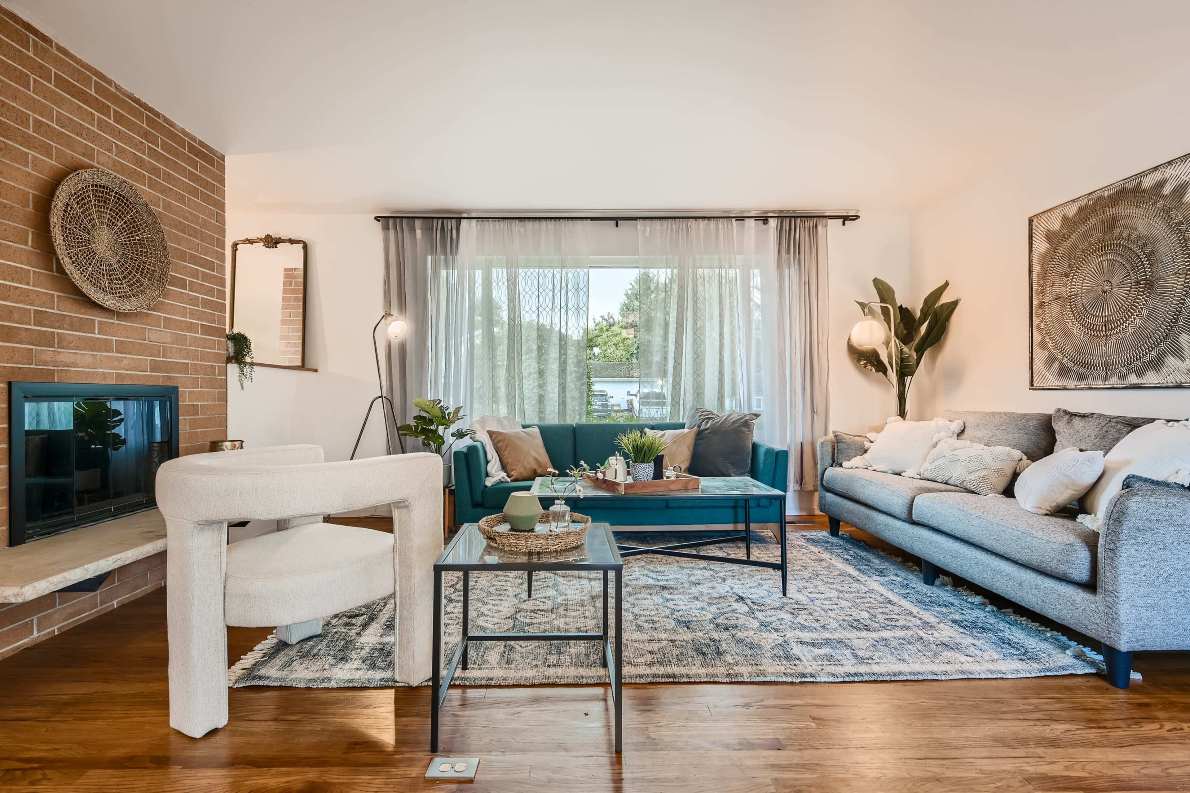 The image shows a living room with a teal sofa, a gray couch, a wooden coffee table, and large windows letting in natural light.