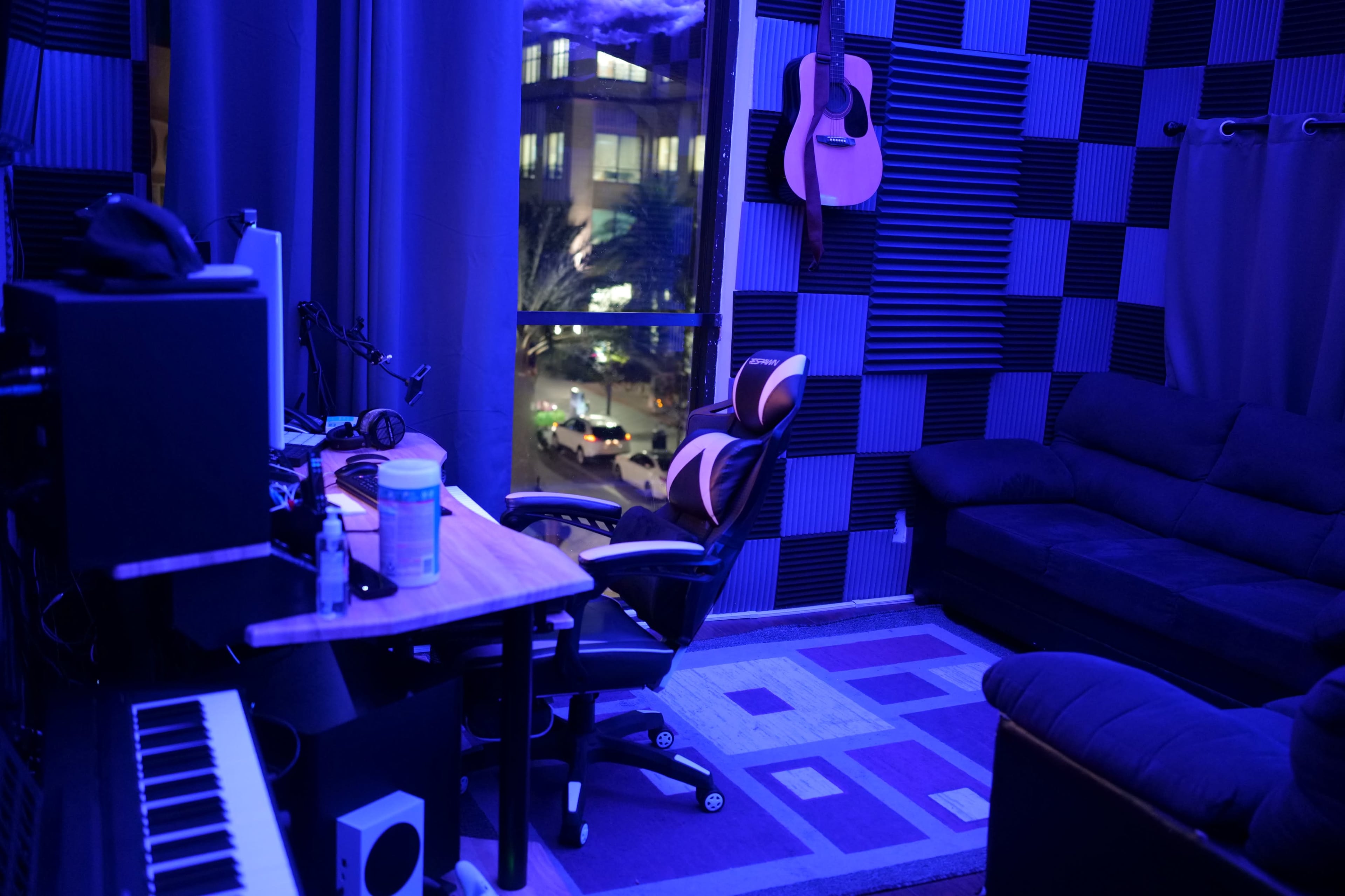 A dimly lit music studio with blue-tinted walls, a desk with computer equipment, a gaming chair, and a sofa, all adjacent to a window revealing a cityscape at night.