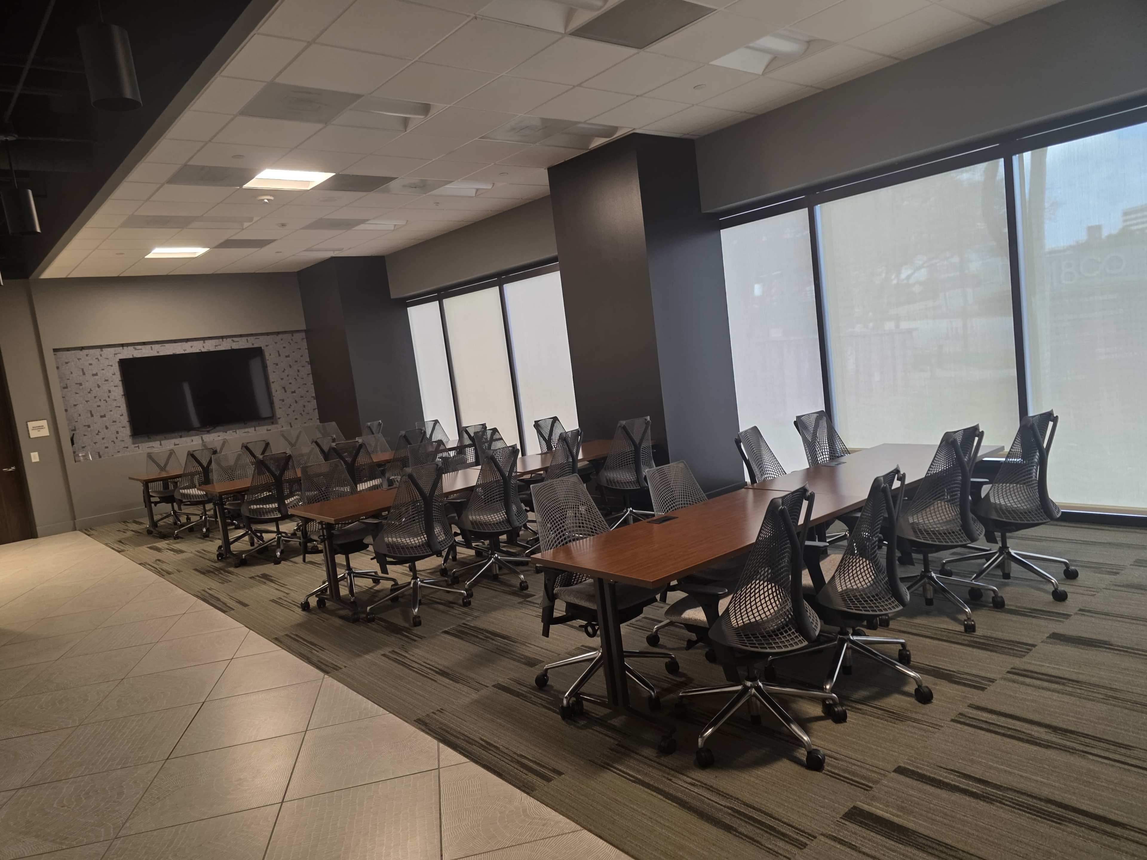 The image shows a modern conference room with multiple tables and ergonomic chairs arranged in rows, large windows allowing natural light to enter, and a wall-mounted television.