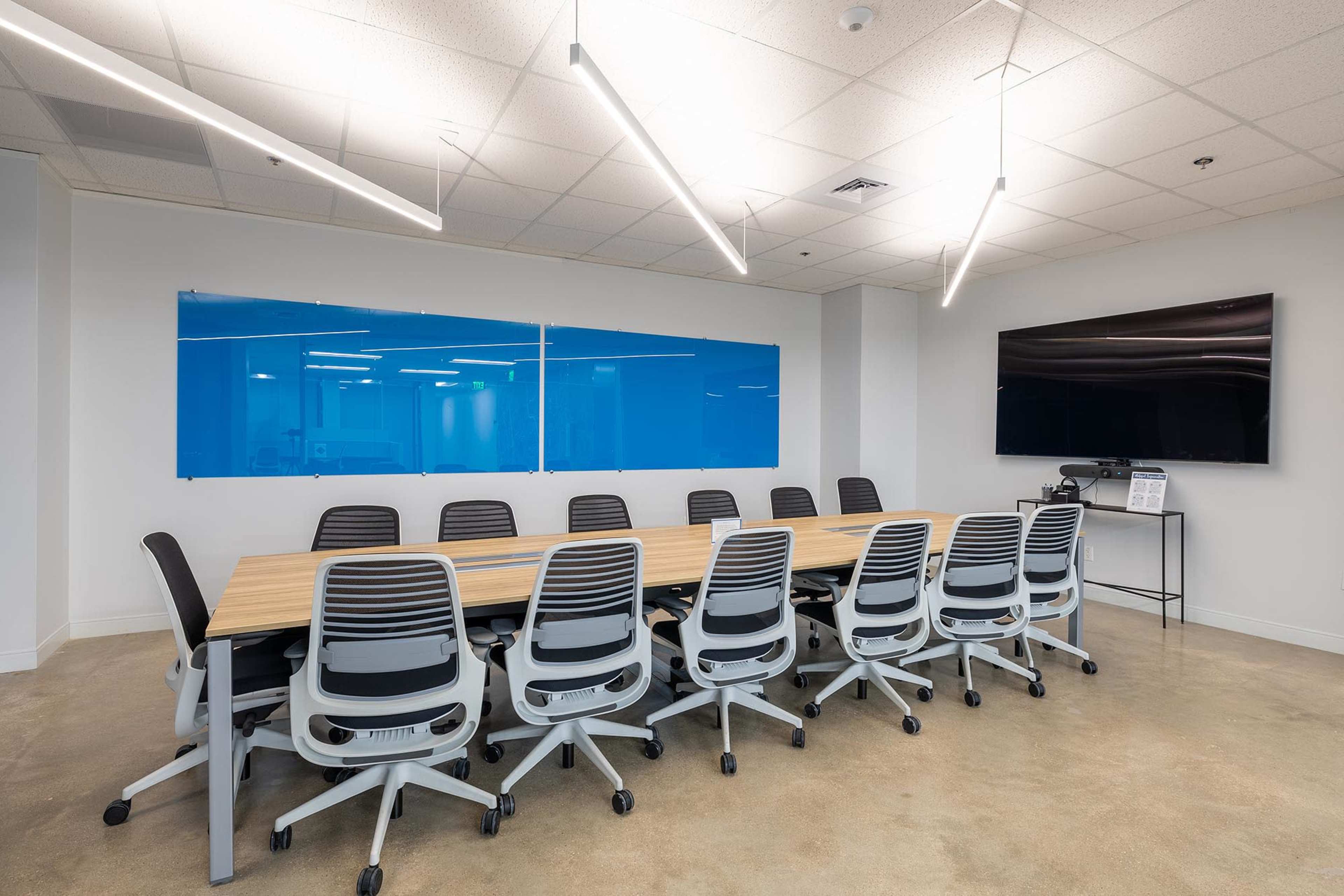 A modern conference room features a long wooden table surrounded by ergonomic swivel chairs, with a large screen mounted on the wall and blue glass panels in the background.
