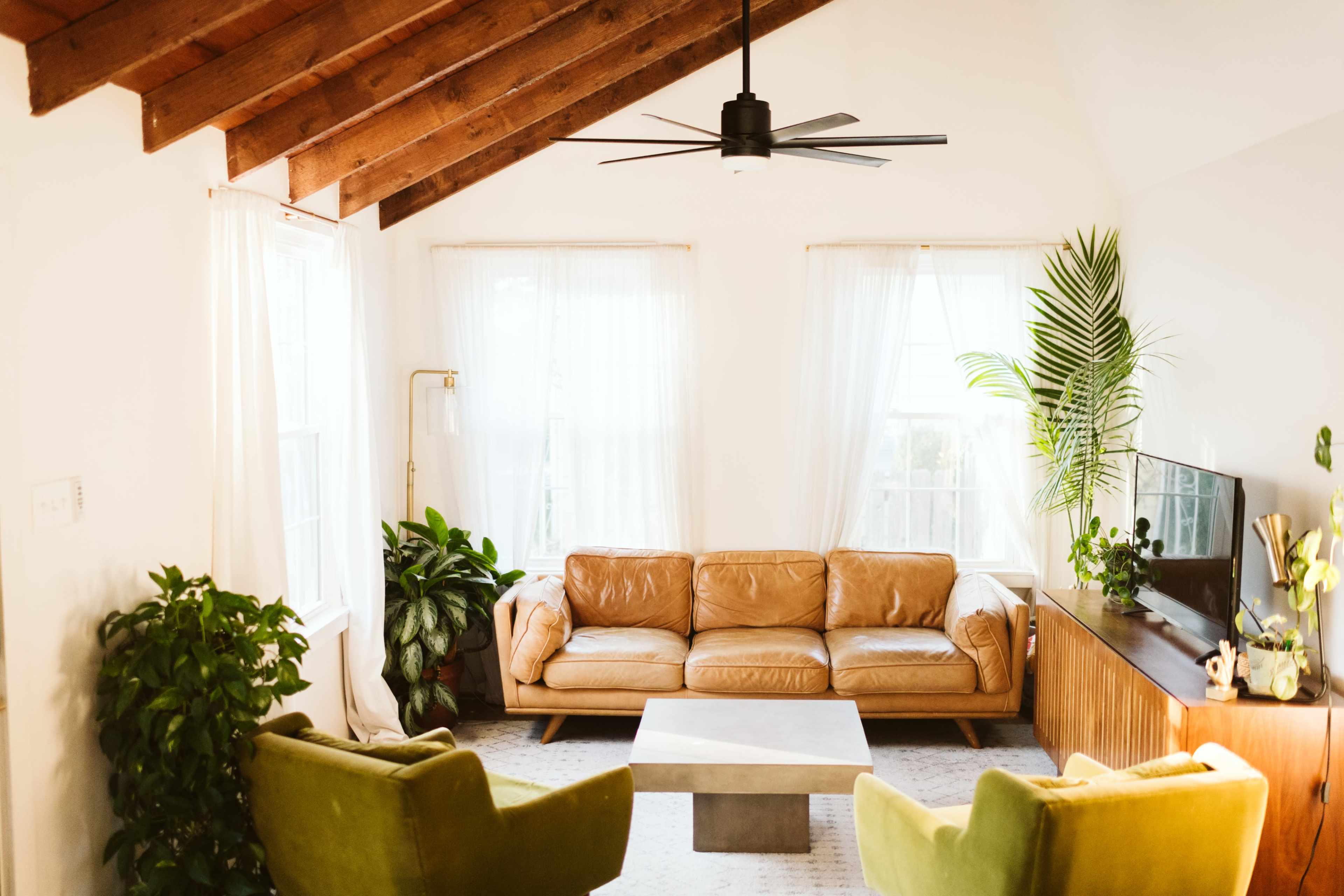 A modern living room features a brown leather sofa, two green armchairs, a coffee table, and large windows with sheer curtains.