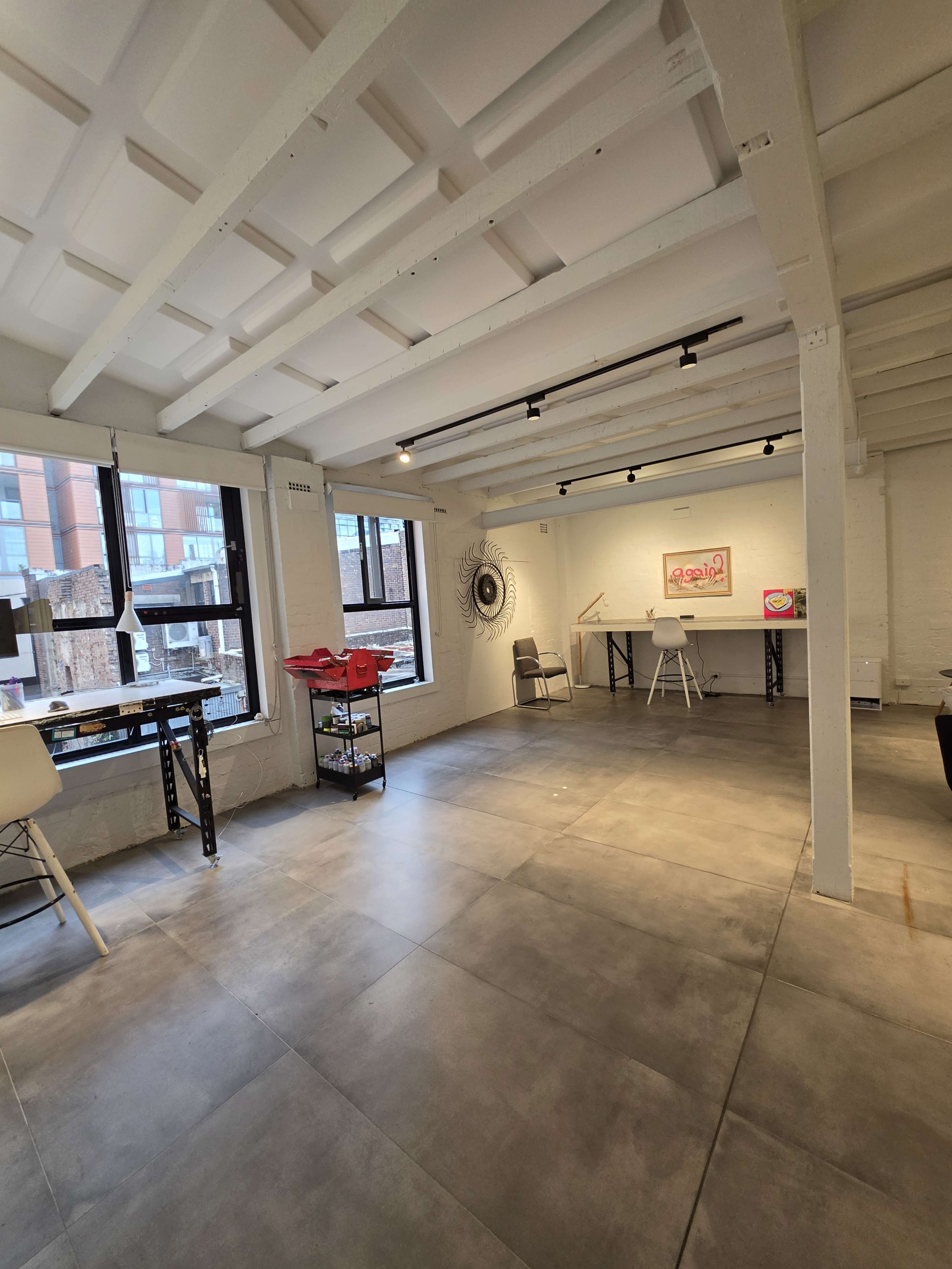 The image shows a bright, modern workspace with white beams, large windows, and minimal furniture.