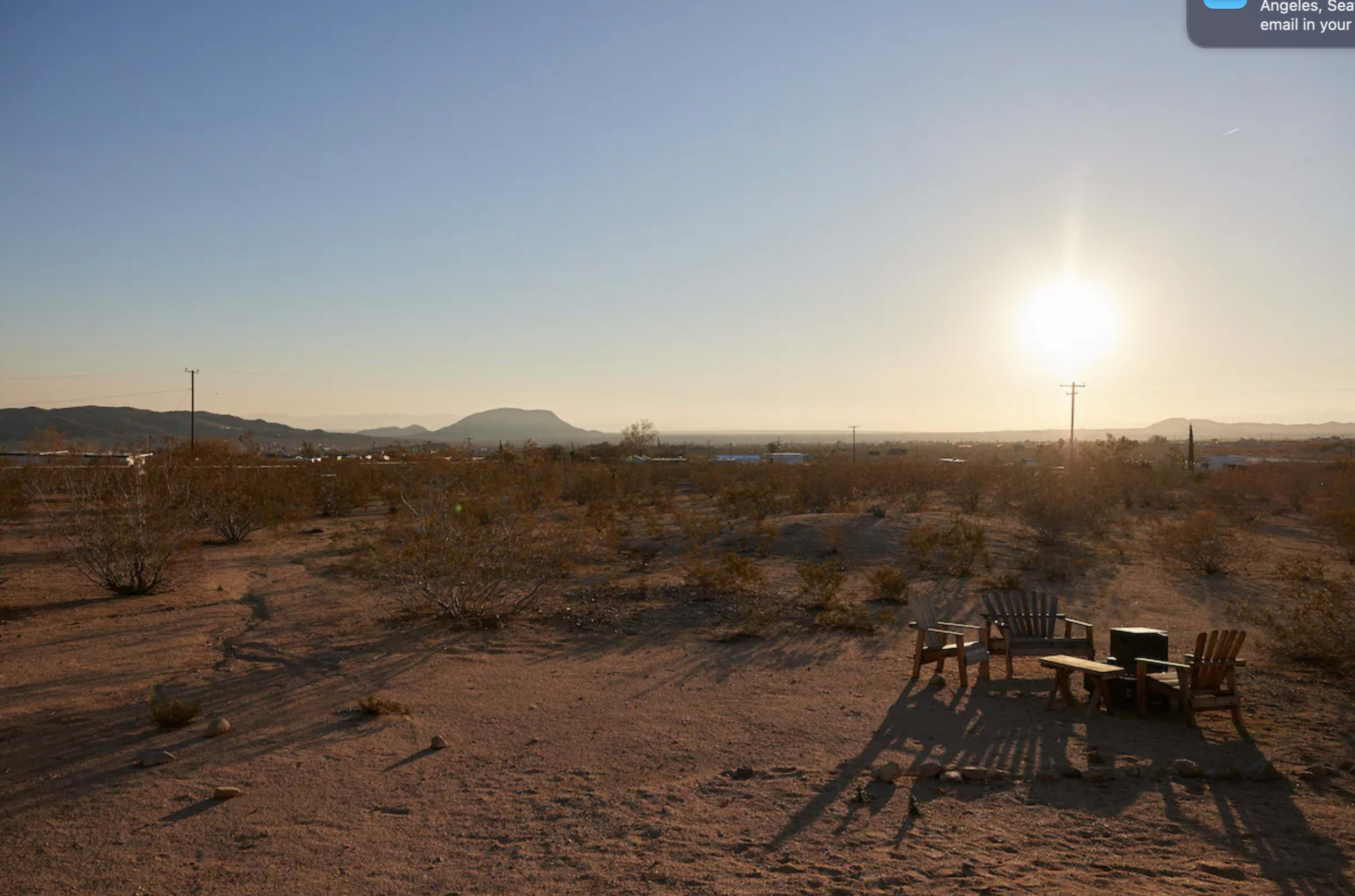 The image shows a desert landscape with a sun setting behind distant mountains, featuring a few wooden chairs arranged on the sandy ground.