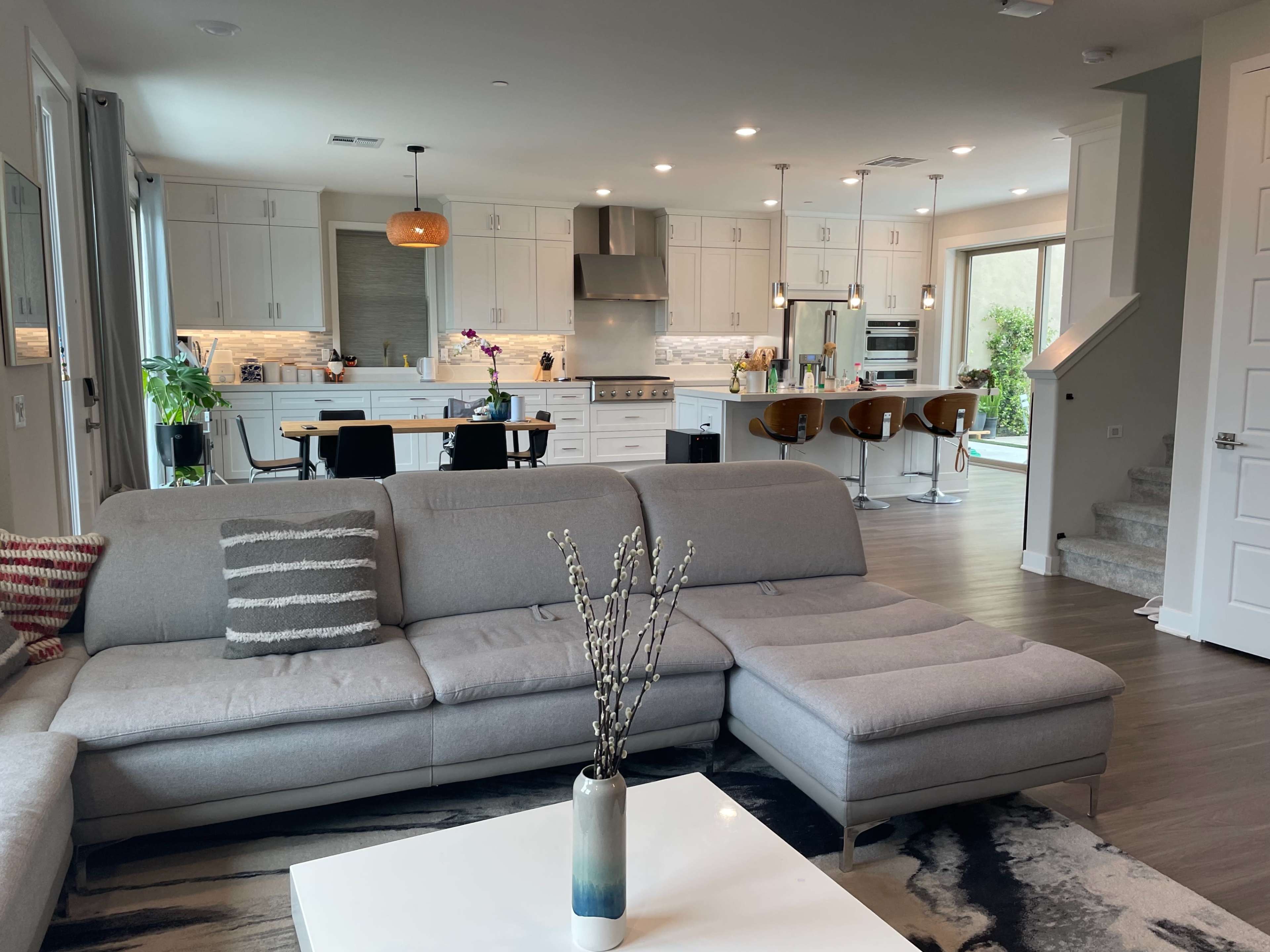 A modern living room connected to a spacious kitchen, featuring a gray sectional sofa, a coffee table with a vase, and large windows allowing natural light.